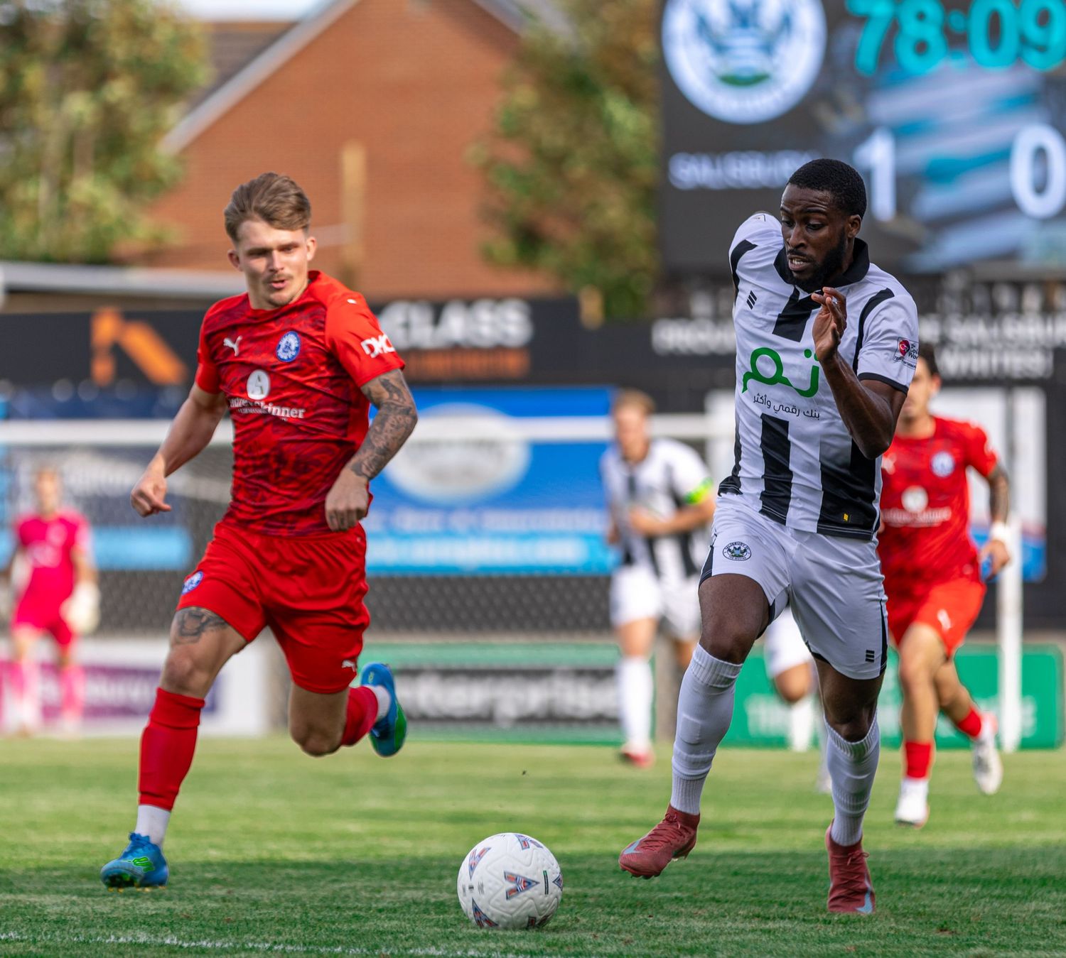 Two soccer players compete for the ball during a match, one in red and one in black and white kit, with a scoreboard showing 78:09.