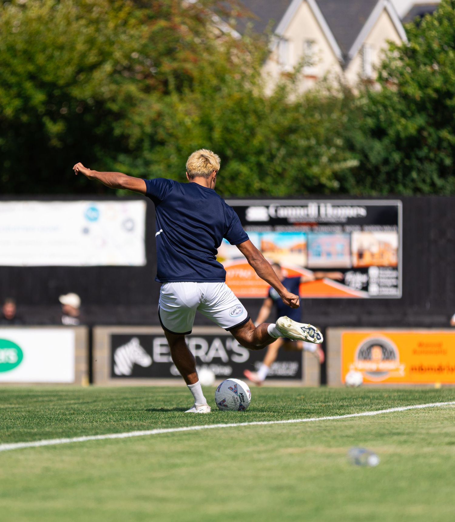 A soccer player in navy blue kit kicks a ball on a green pitch during a sunny outdoor match.