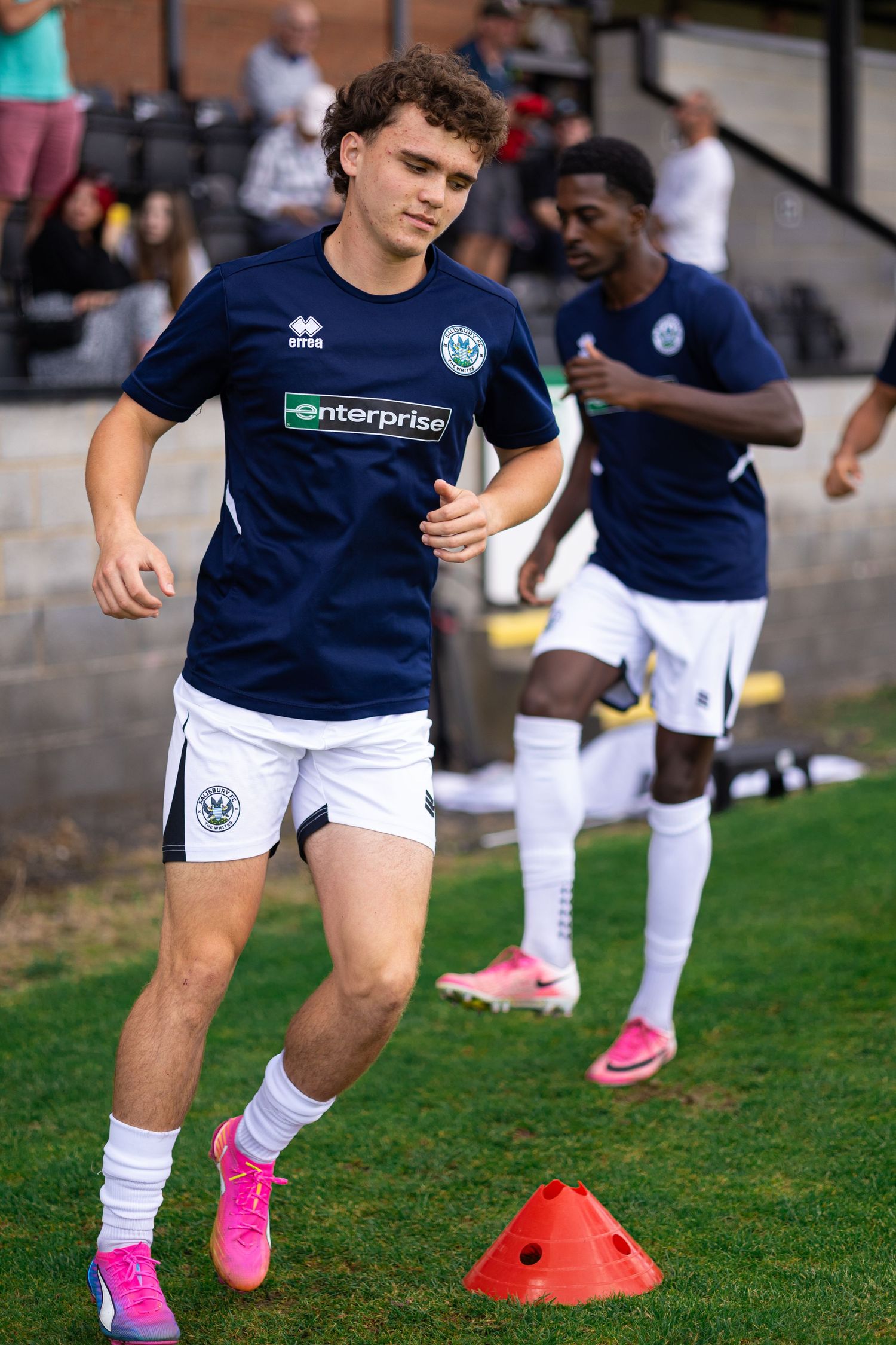 Soccer player in navy Enterprise-sponsored jersey and white shorts dribbles past orange cone during warm-up drill.