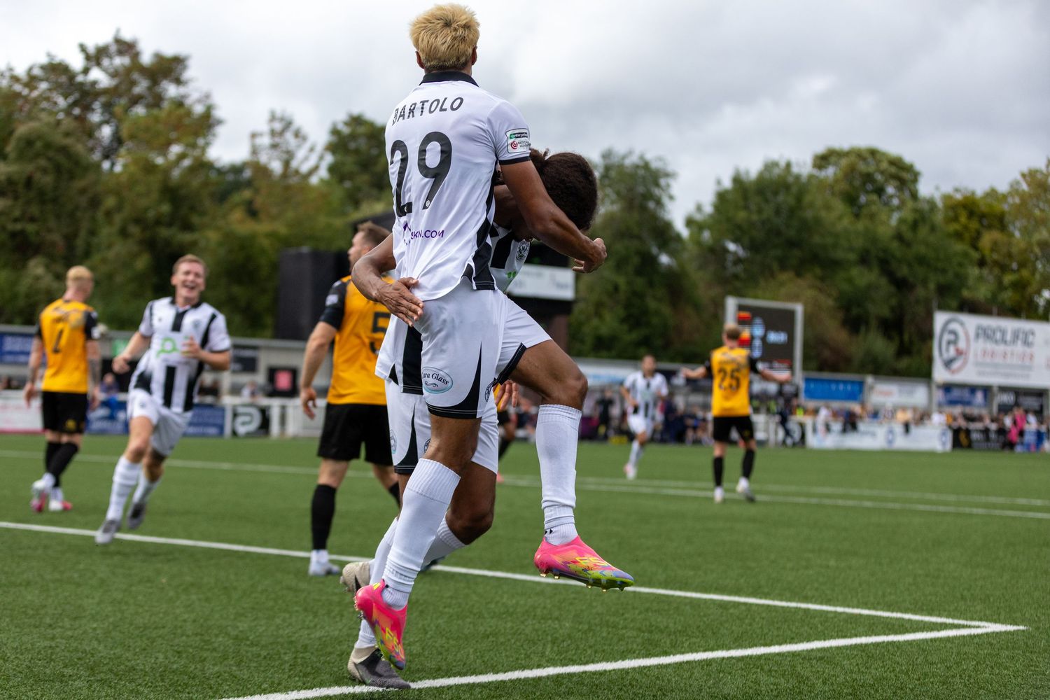 Two soccer players in white jerseys celebrate a goal during a match, with number 29 jumping on teammate's back.