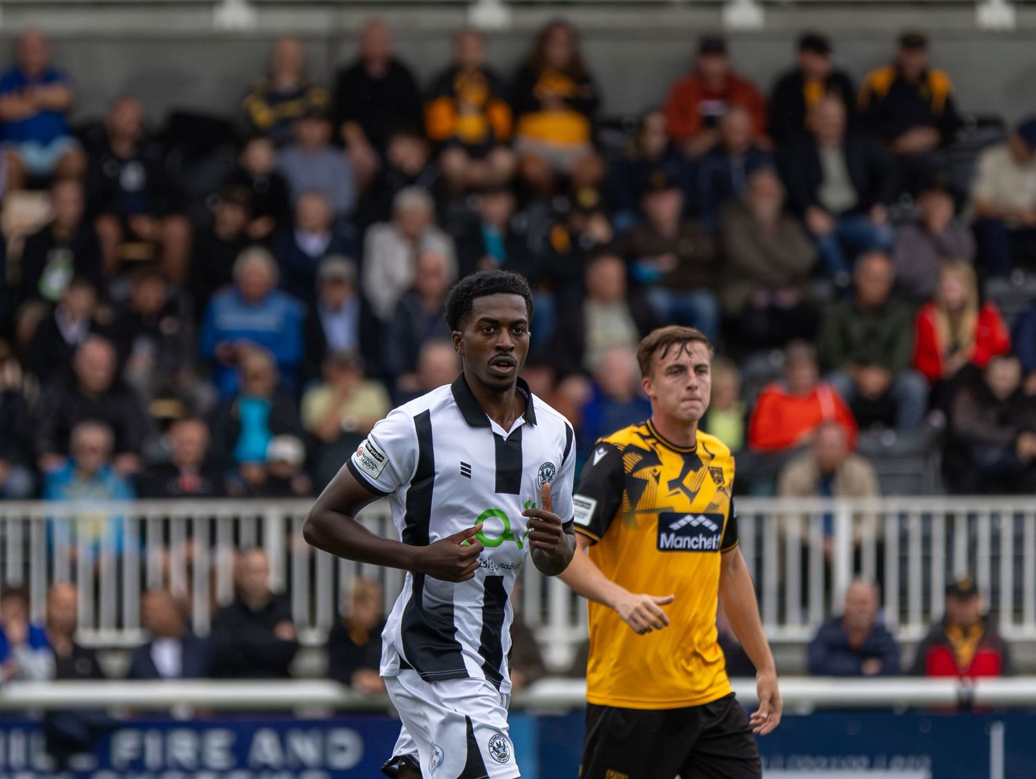 Two soccer players in contrasting black-and-white and yellow jerseys compete on a football pitch with spectators in the background.
