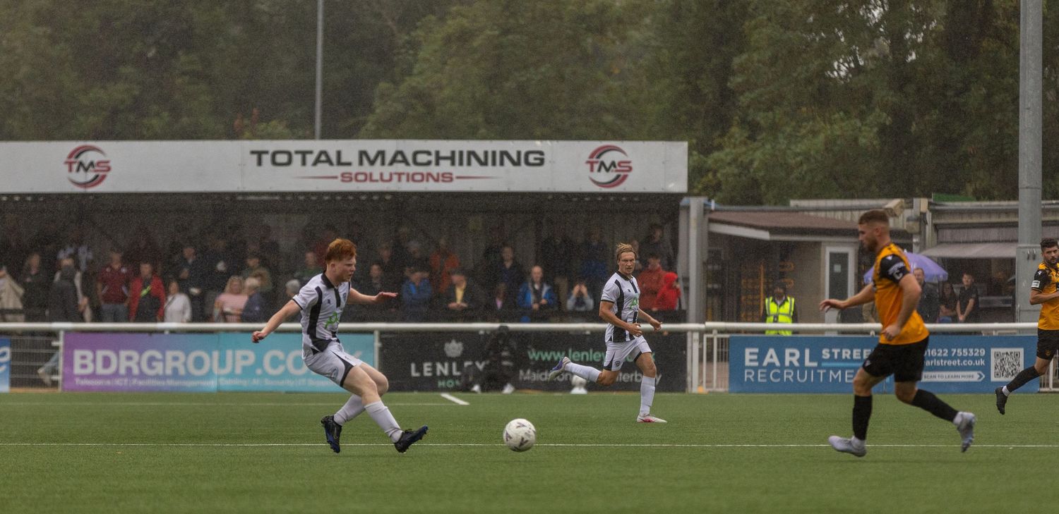 Football players compete on a pitch with Total Machining Solutions advertising boards visible in the background.