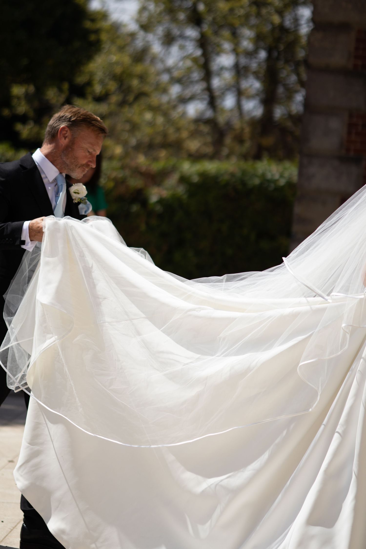 Groom in black suit holding flowing white wedding dress train outdoors on sunny day.