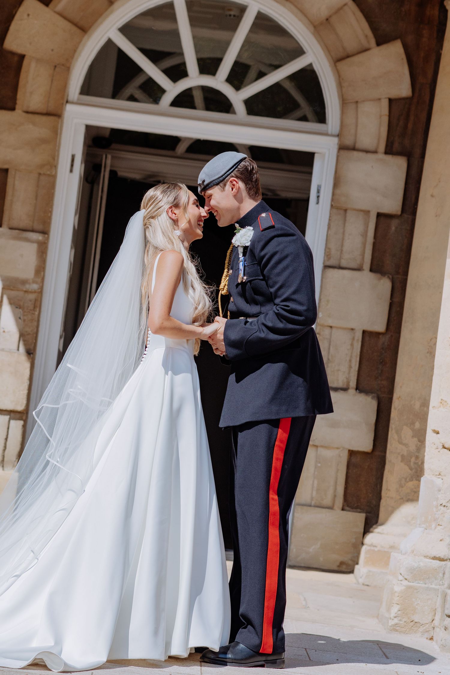 Newlyweds share an intimate moment outside a stone building, groom wearing military dress uniform.