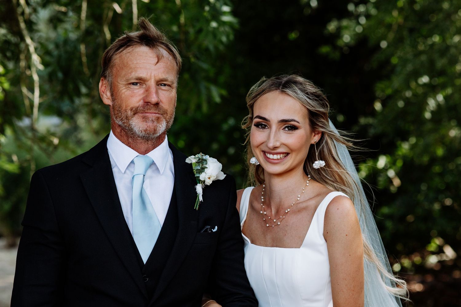 Bride in white wedding dress and veil posing with older gentleman in black suit and blue tie.