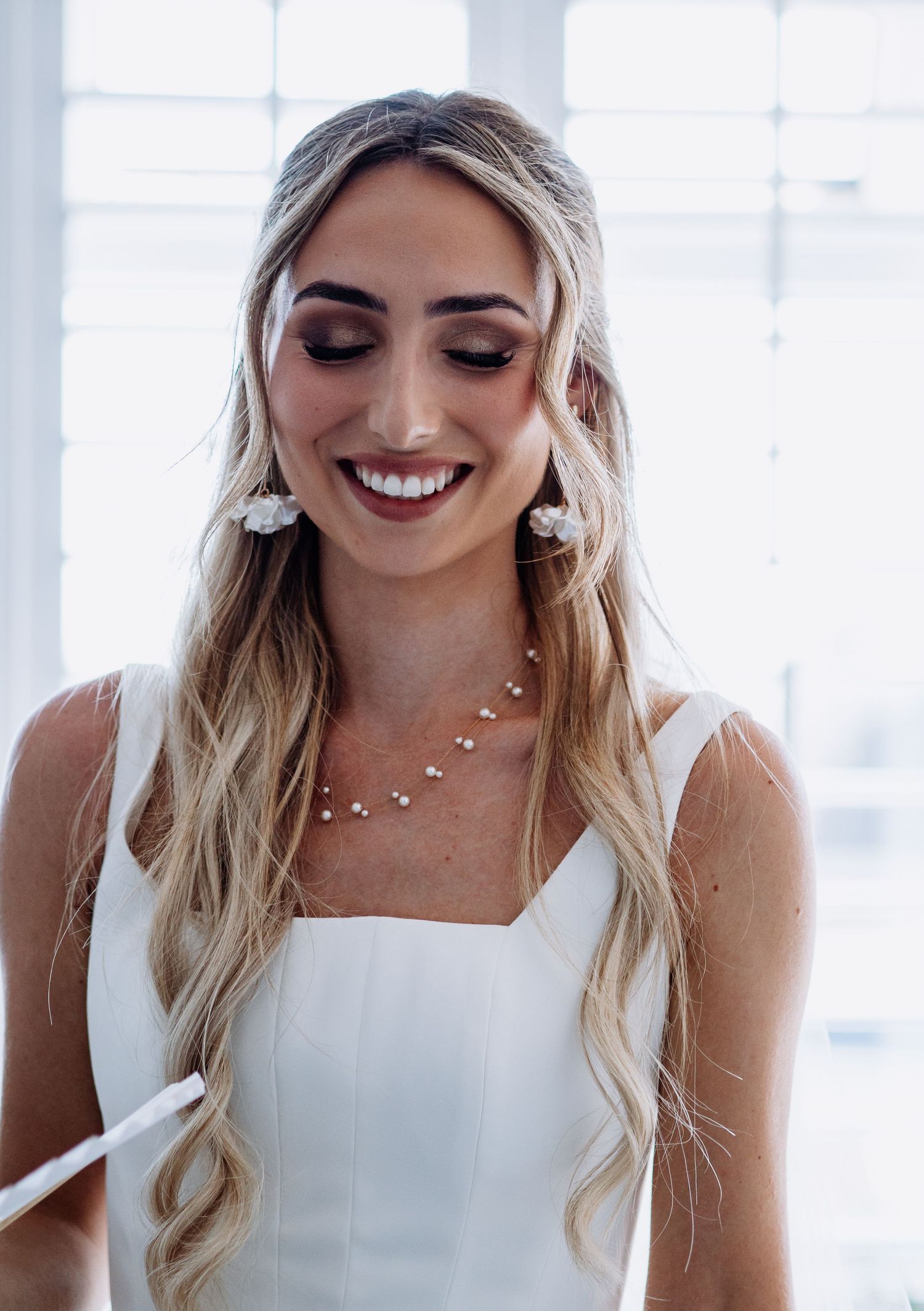 Beautiful bride in white dress with long blonde waves smiling downward wearing floral earrings and necklace.
