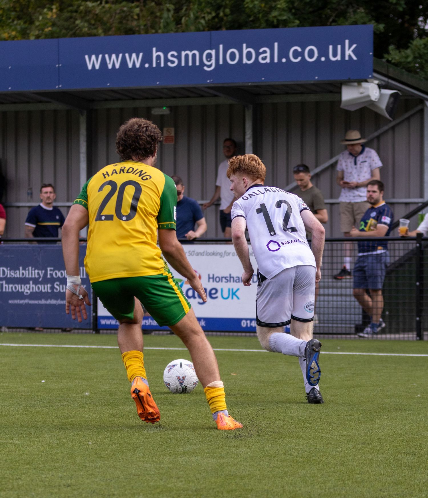 Gallagher number 12 in white challenges Harding number 20 in yellow kit during an outdoor football match.