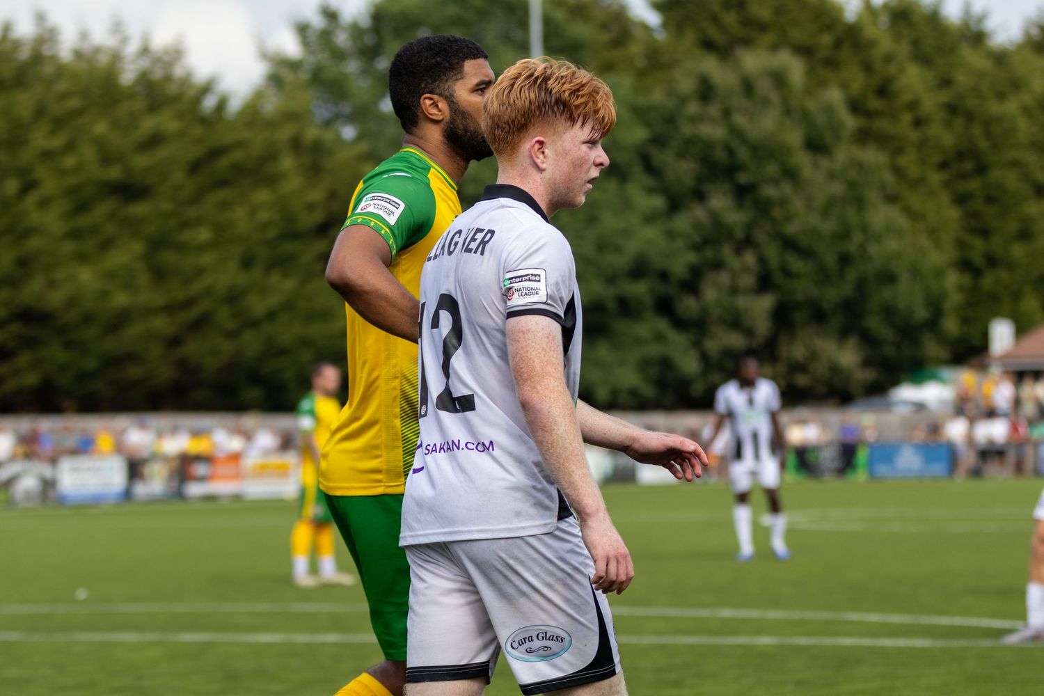 Footballer in yellow-green kit walks closely behind white number 2 jersey player during outdoor match.