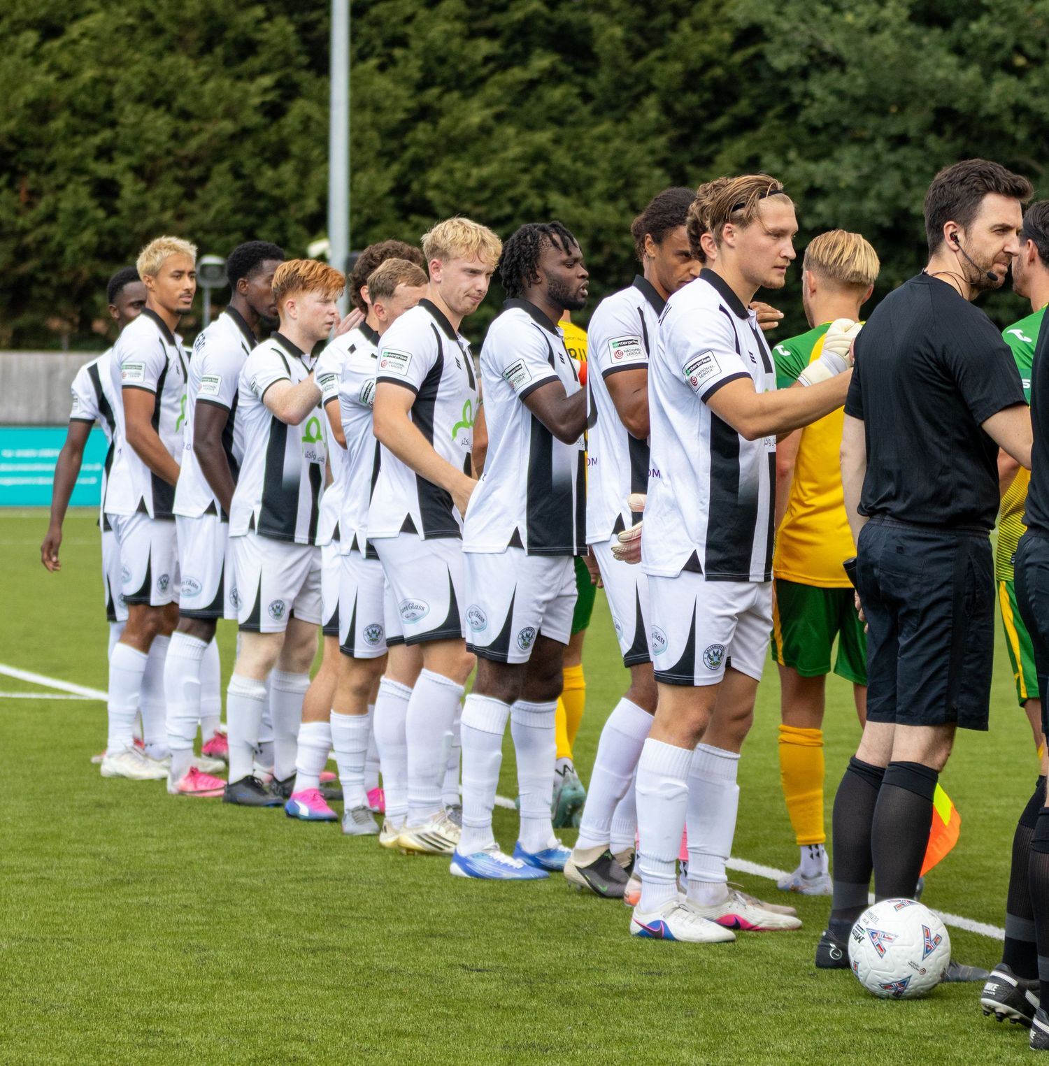 A football team in white kits lines up on the pitch before a non-league match on a sunny day.