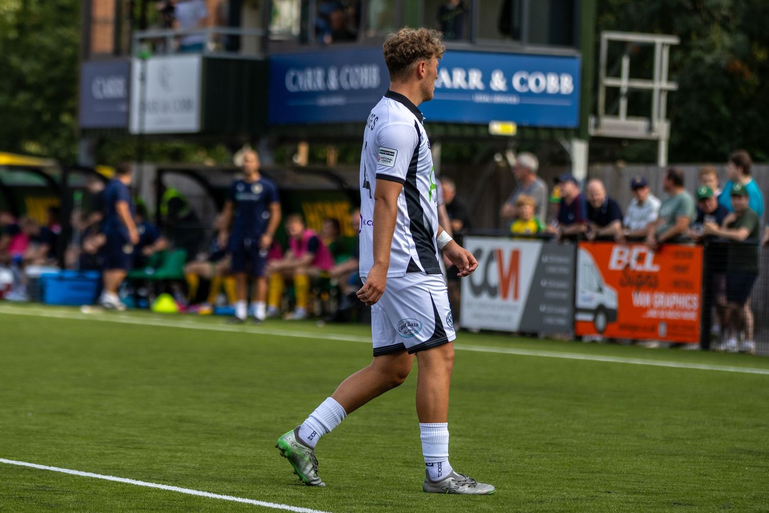 A soccer player in white kit walks along the sideline during a match at Carr & Cobb sponsored ground.