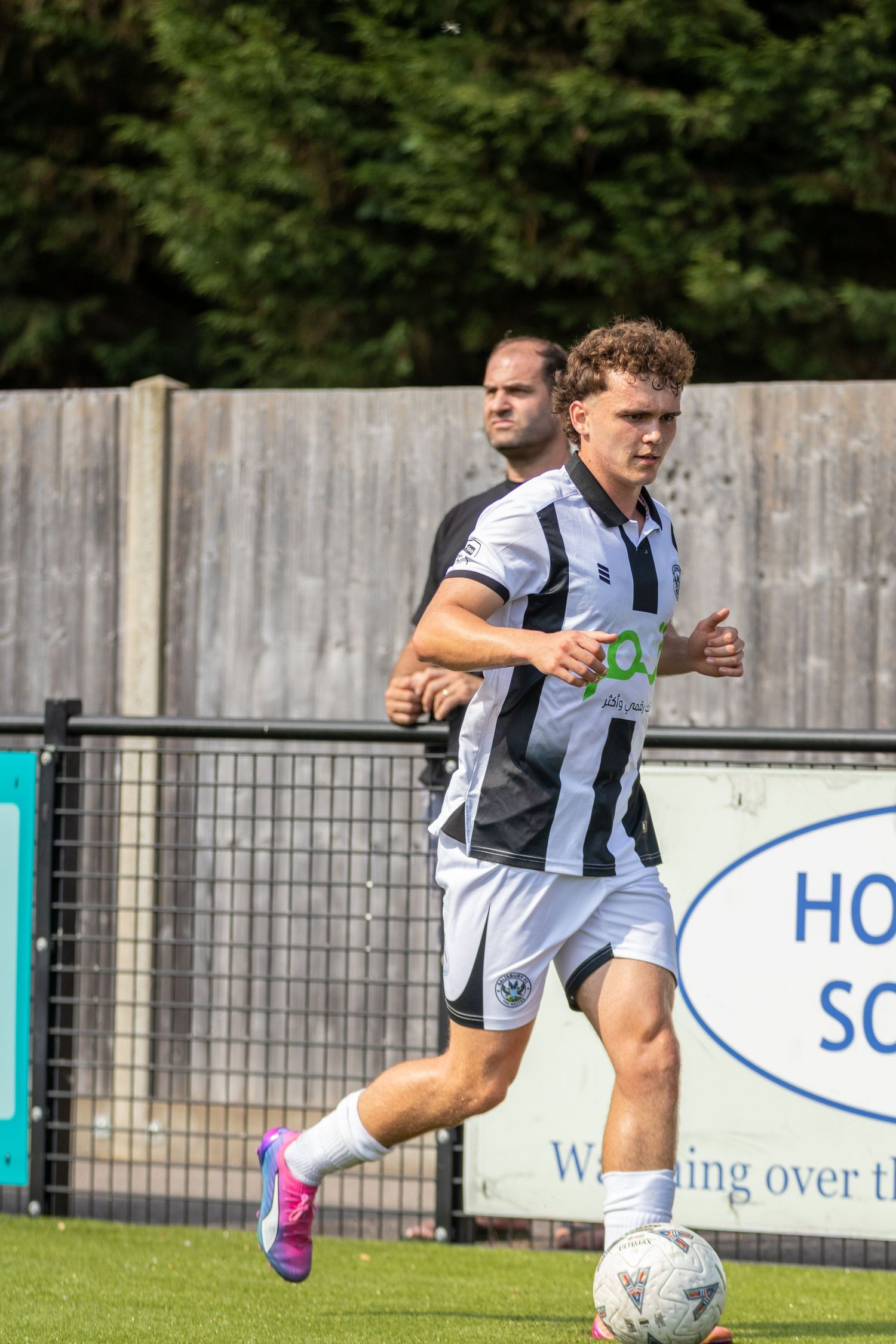 A curly-haired footballer in black and white kit runs with the ball during an outdoor football match.