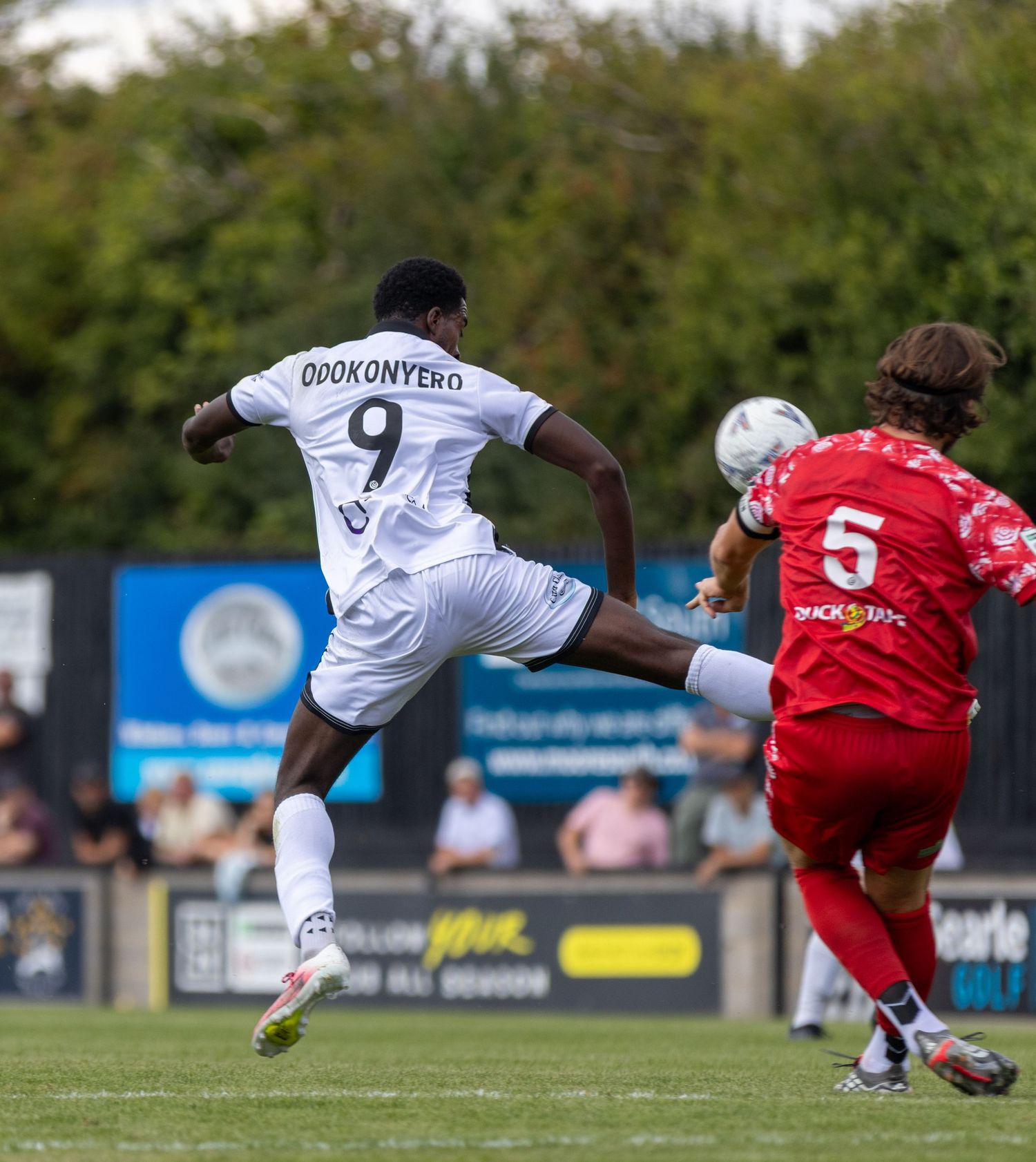 Odokonyero number 9 in white kit battles for the ball against a red-kitted defender number 5 in a football match.