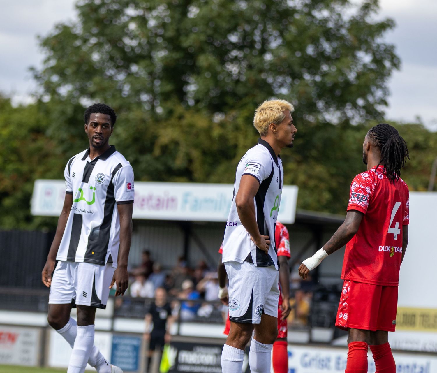 Football players in black-white and red kits stand on the pitch during a pause in play.