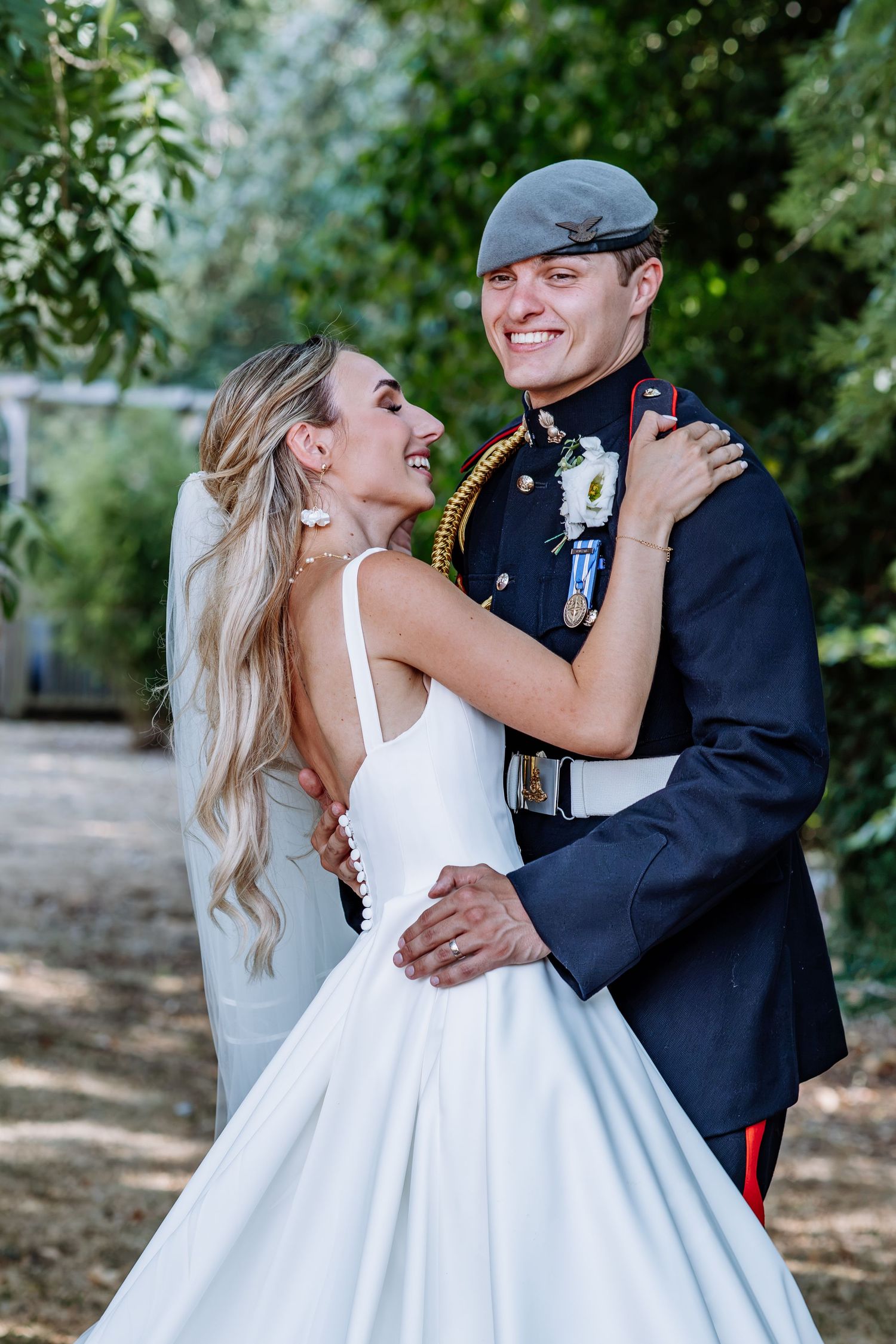 Smiling bride and groom in military dress uniform embracing joyfully during outdoor wedding portraits.