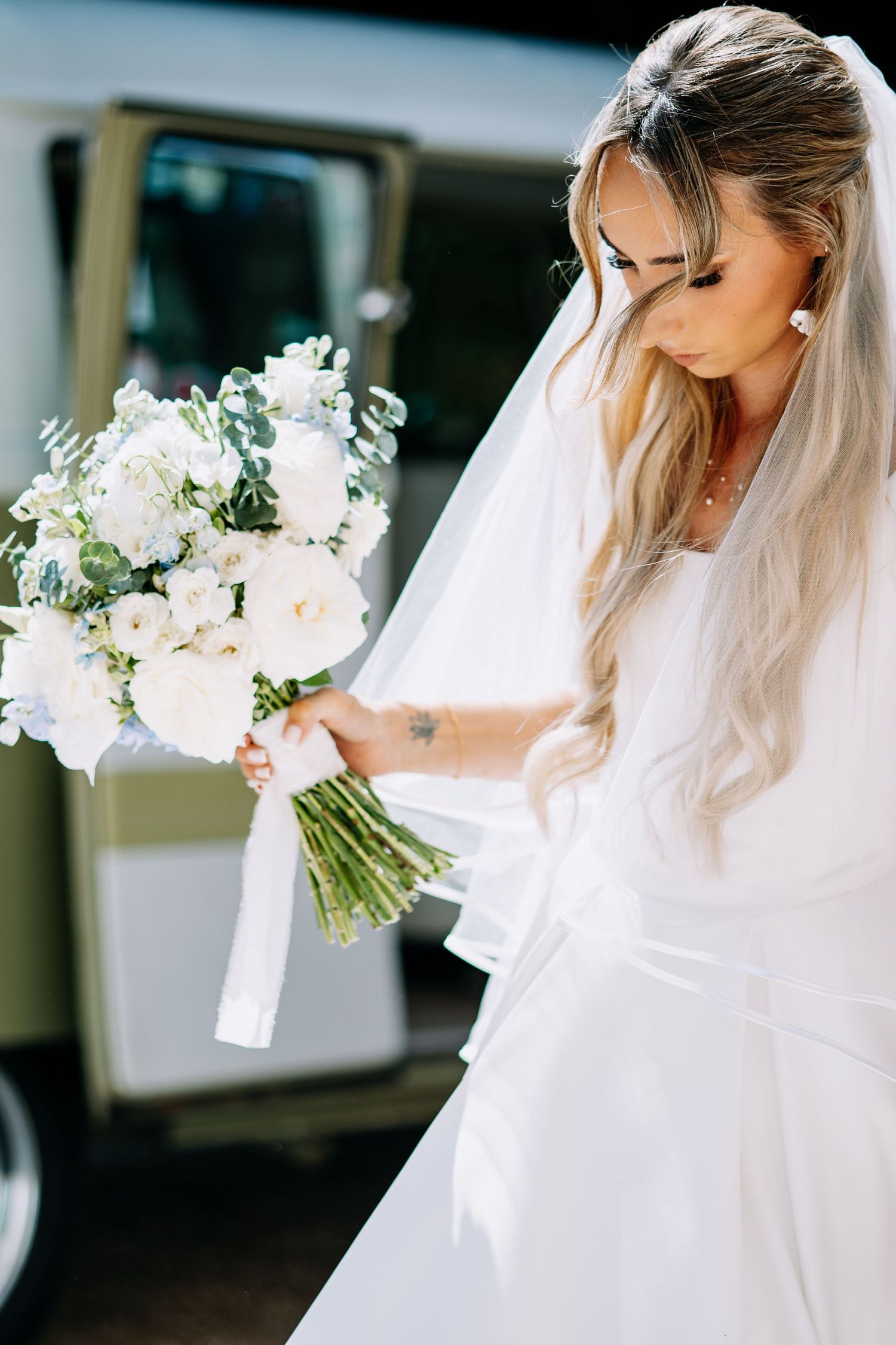 Bride in white satin wedding dress and veil holding white bouquet with ribbon detail.