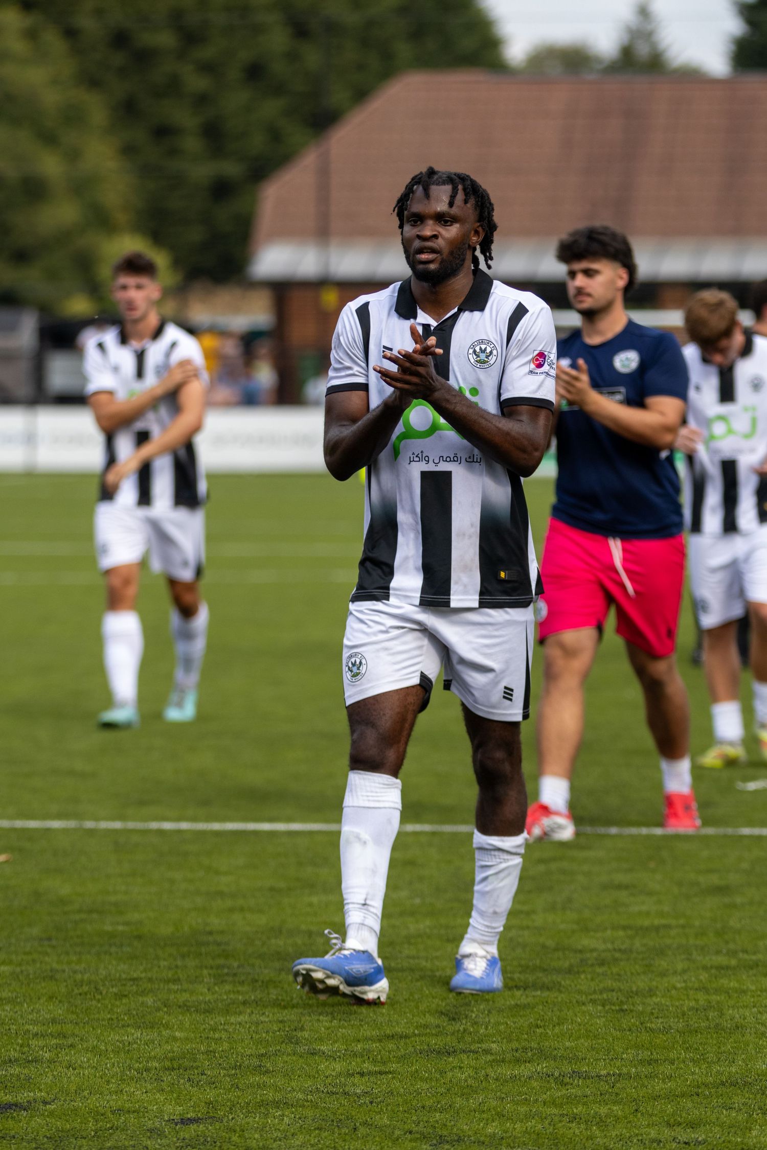 A tall footballer with dreadlocks in black and white kit applauds supporters after a football match.