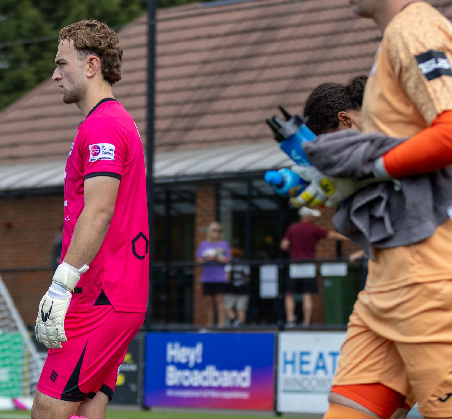 A goalkeeper in a pink kit stands beside another goalkeeper in orange during a non-league football match.