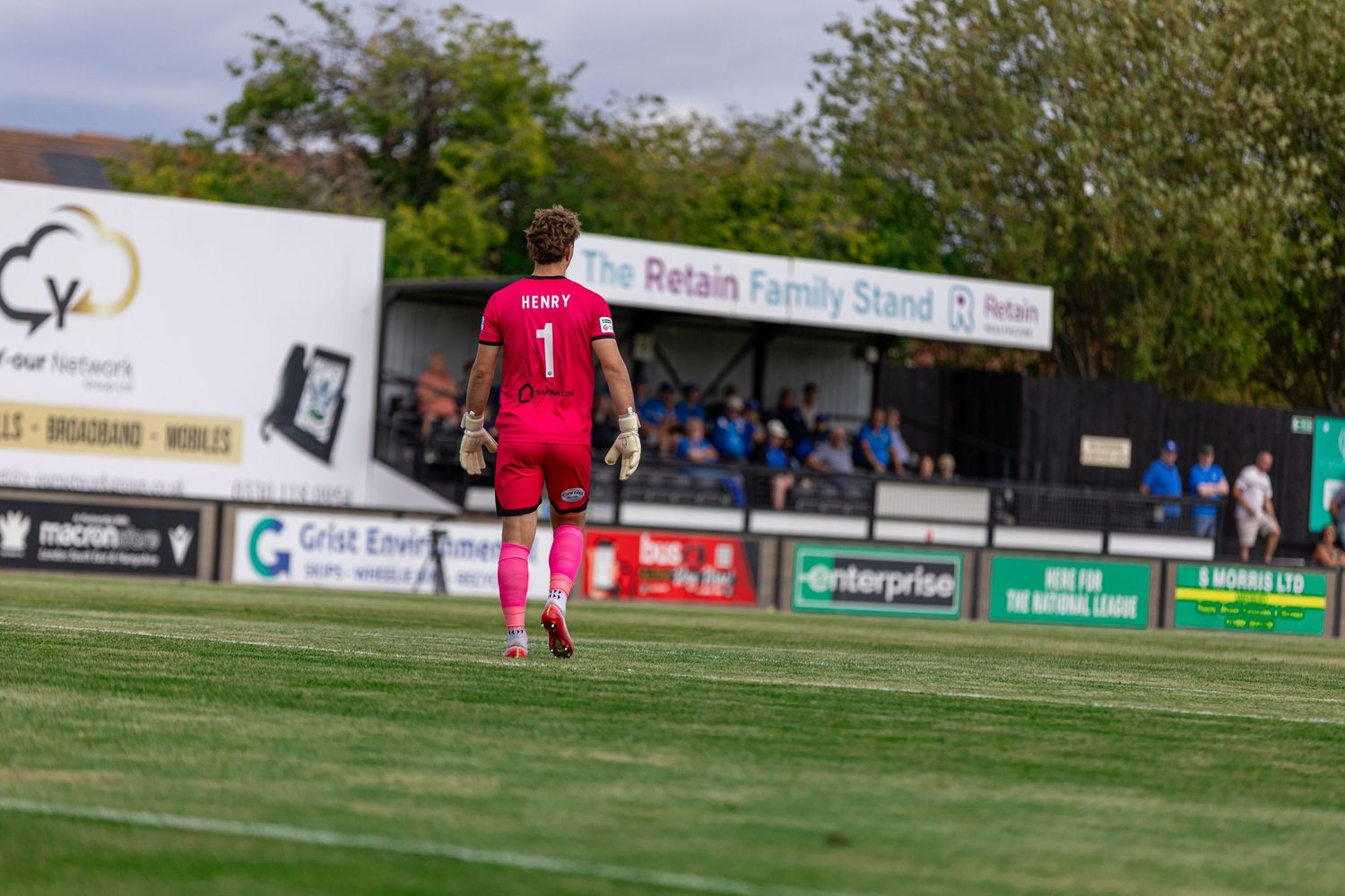 Goalkeeper in red kit walks on green pitch near the Retain Family Stand during a football match.