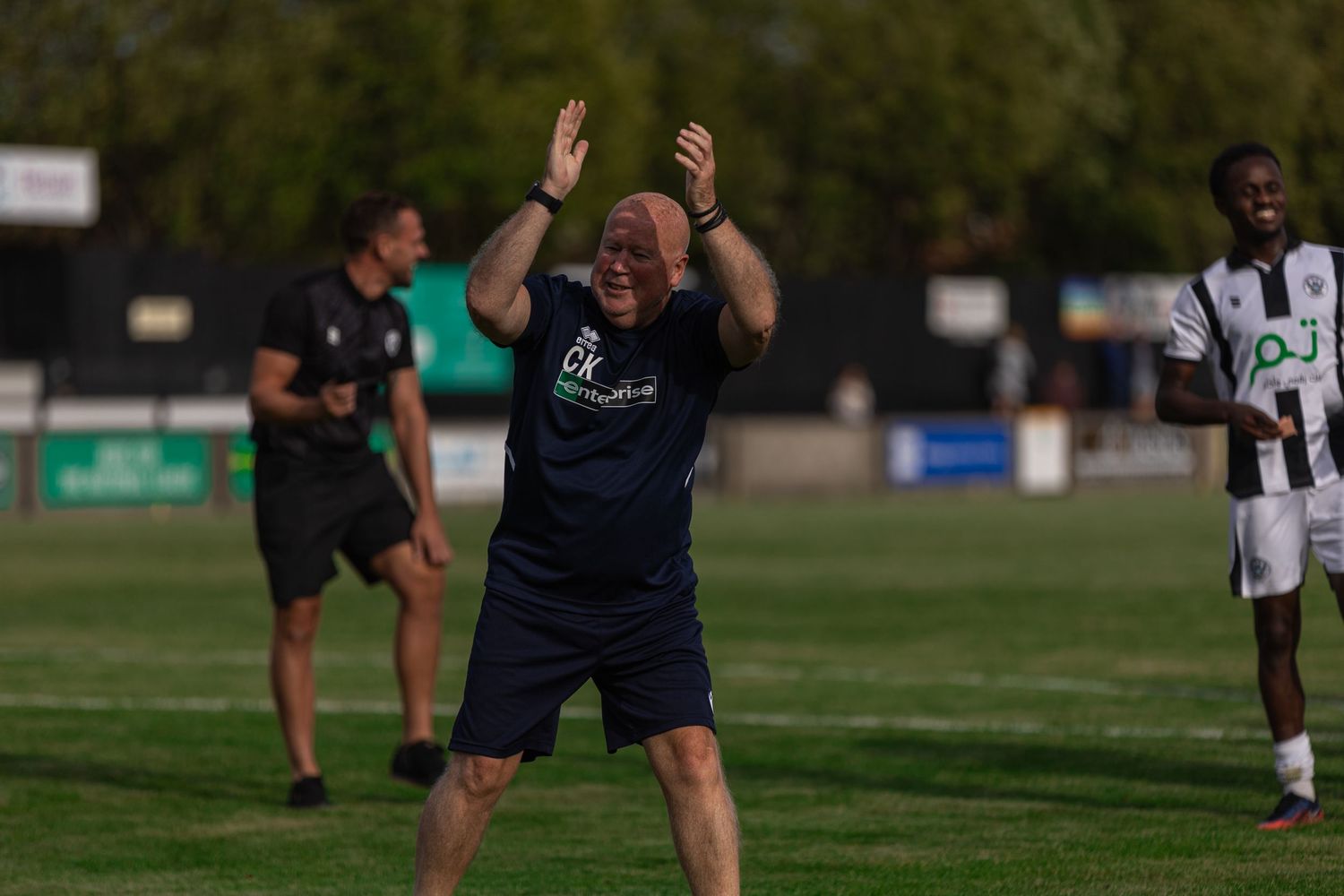 A jubilant football coach celebrates with arms raised on a sunny outdoor pitch during a match.