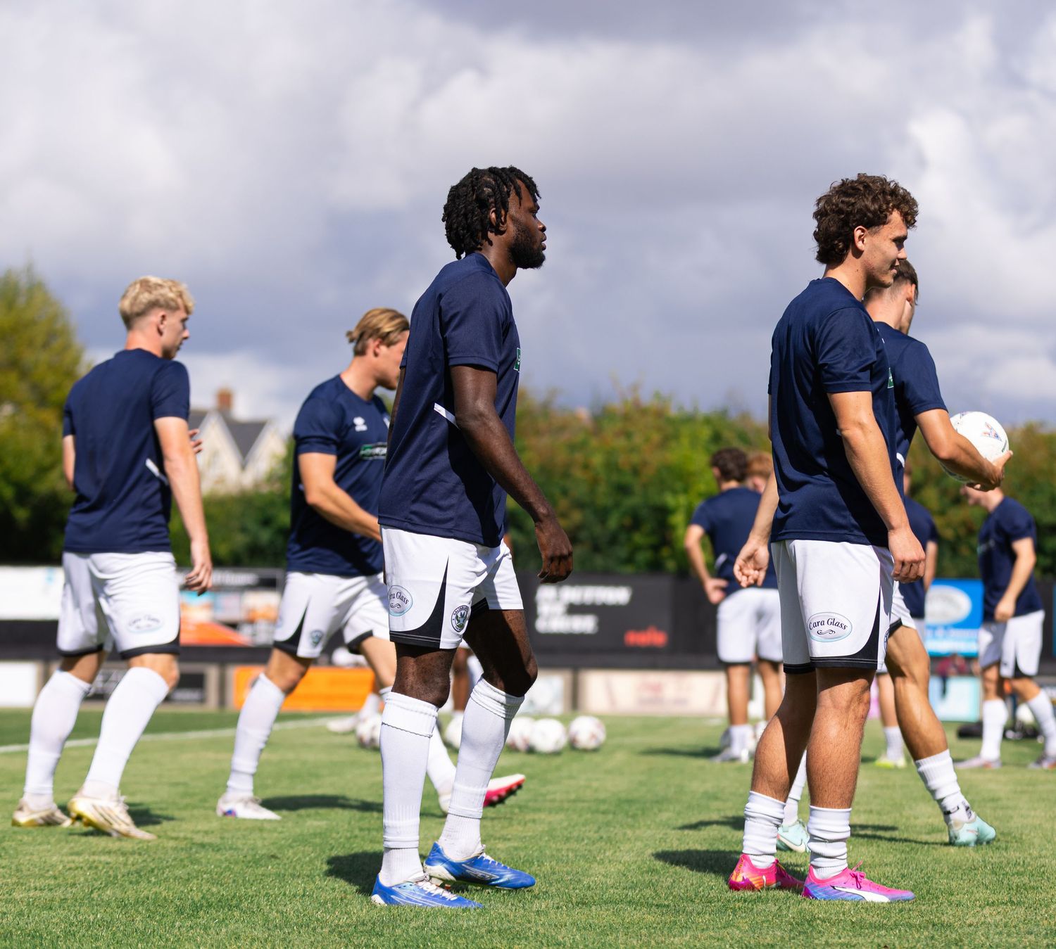 Football players in navy blue kits warming up on a grass pitch before a match on a cloudy day.