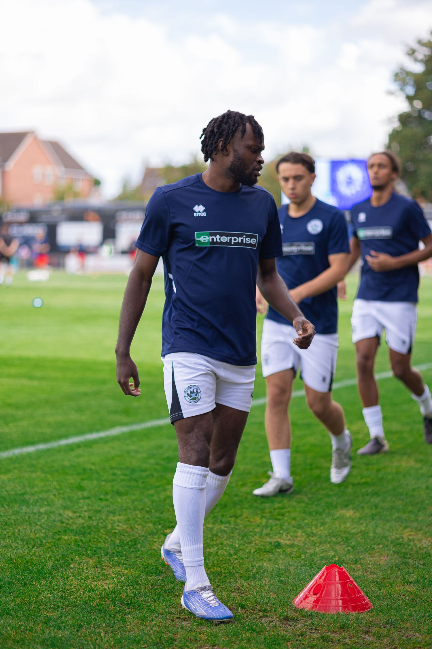 Football players in navy Enterprise-sponsored jerseys warm up on a green pitch with red training cones during pre-match preparation.