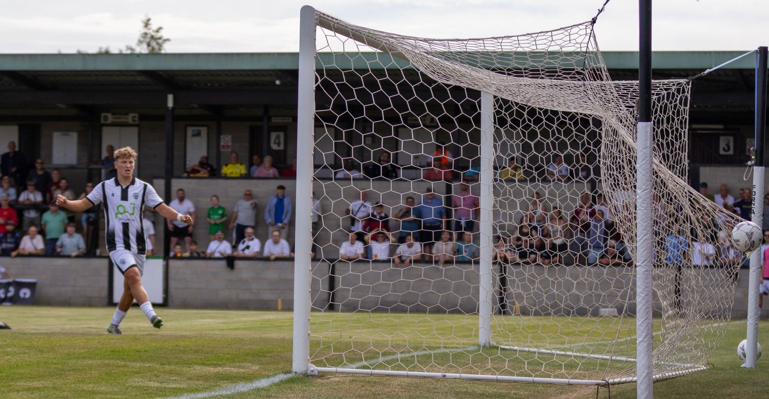 Football player in black and white walks near the goal post after scoring during an outdoor match.
