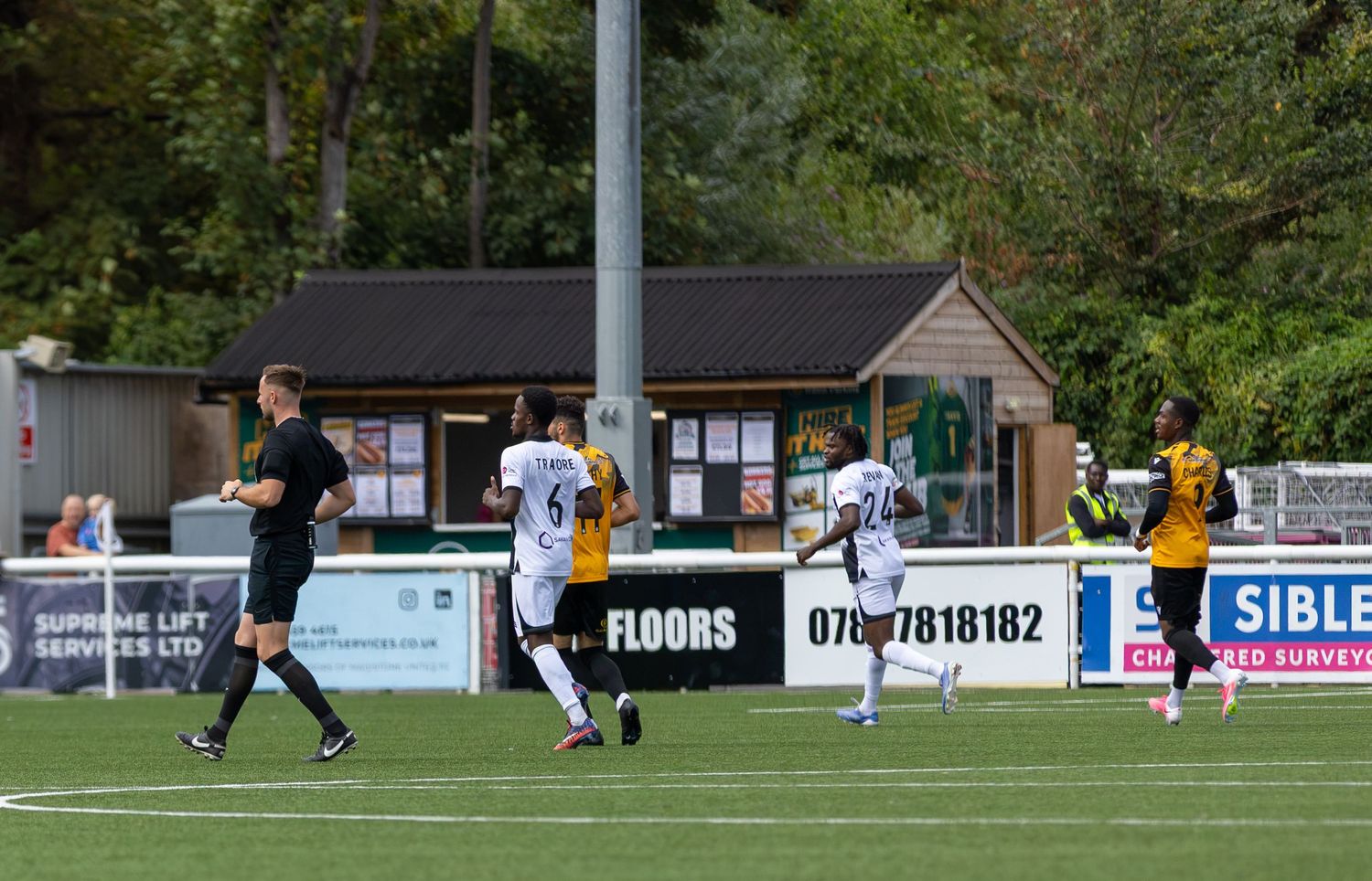 Soccer players compete on a green pitch during a sunny outdoor match, with spectators and trees visible in the background.