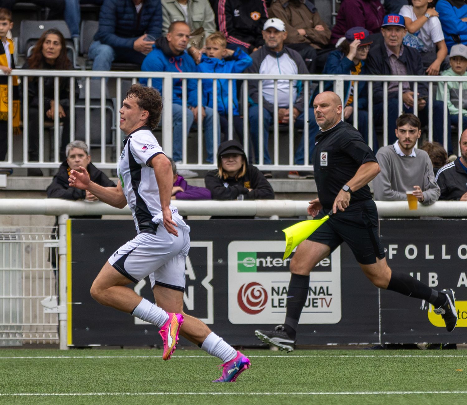 Soccer player in black and white kit sprints past referee during National League TV match at outdoor stadium.