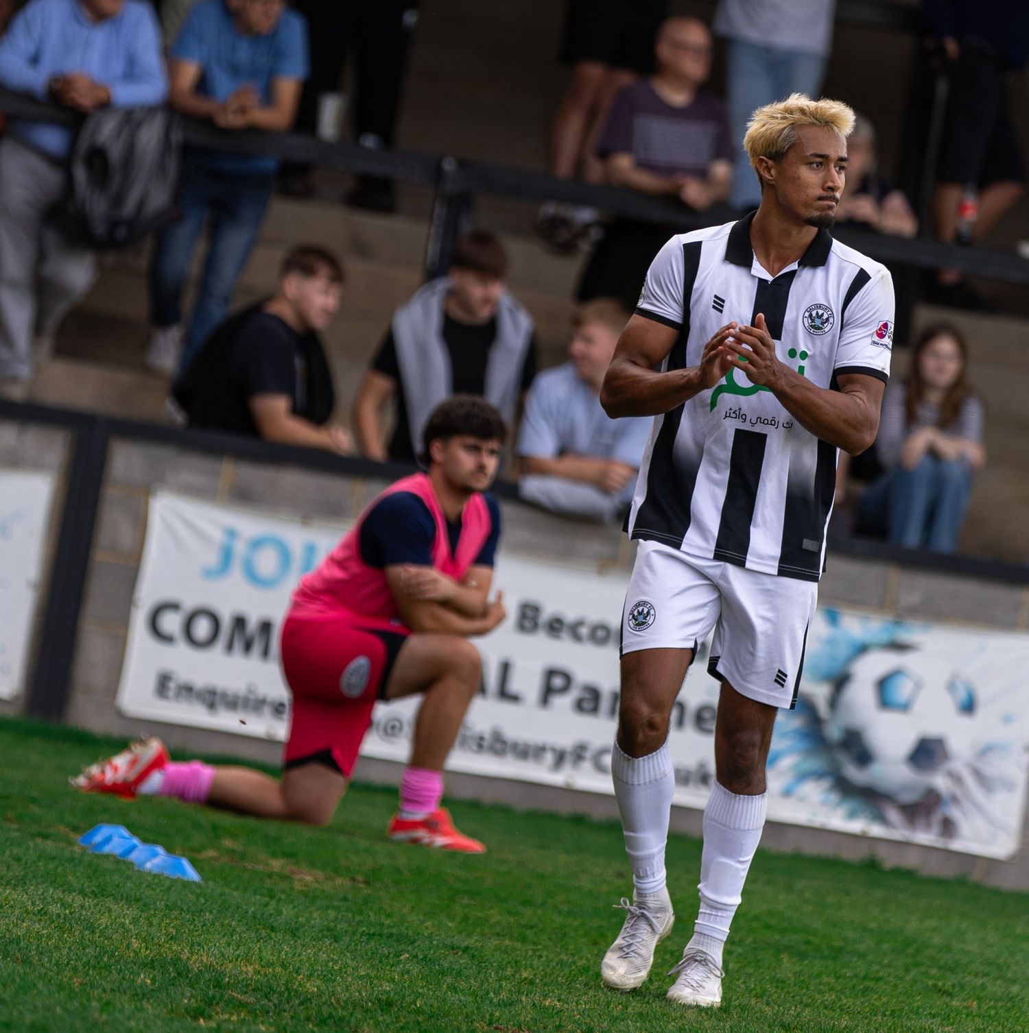 Soccer player in black and white kit holds ball on green pitch, with teammate and spectators visible in background.