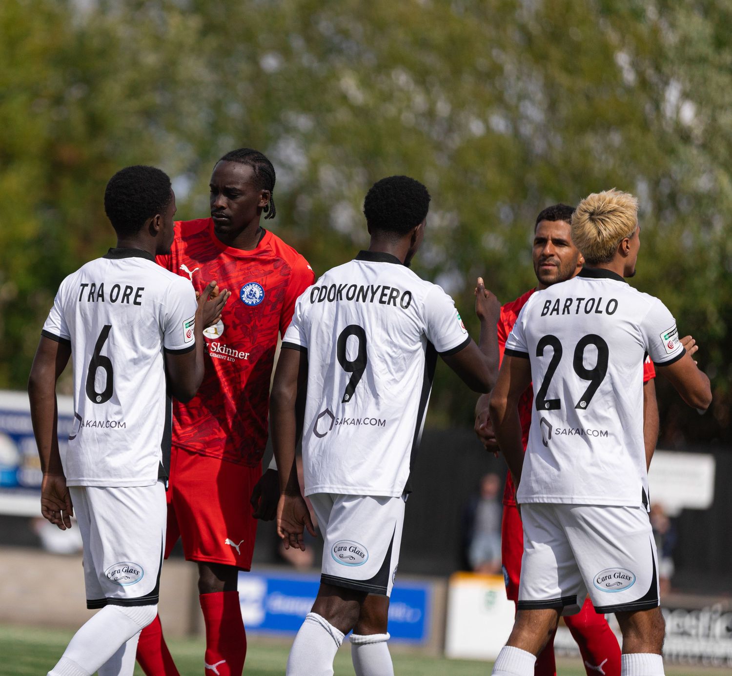 Soccer players in white jerseys with numbers 6, 9, and 29 huddle on field during a match on a sunny day.