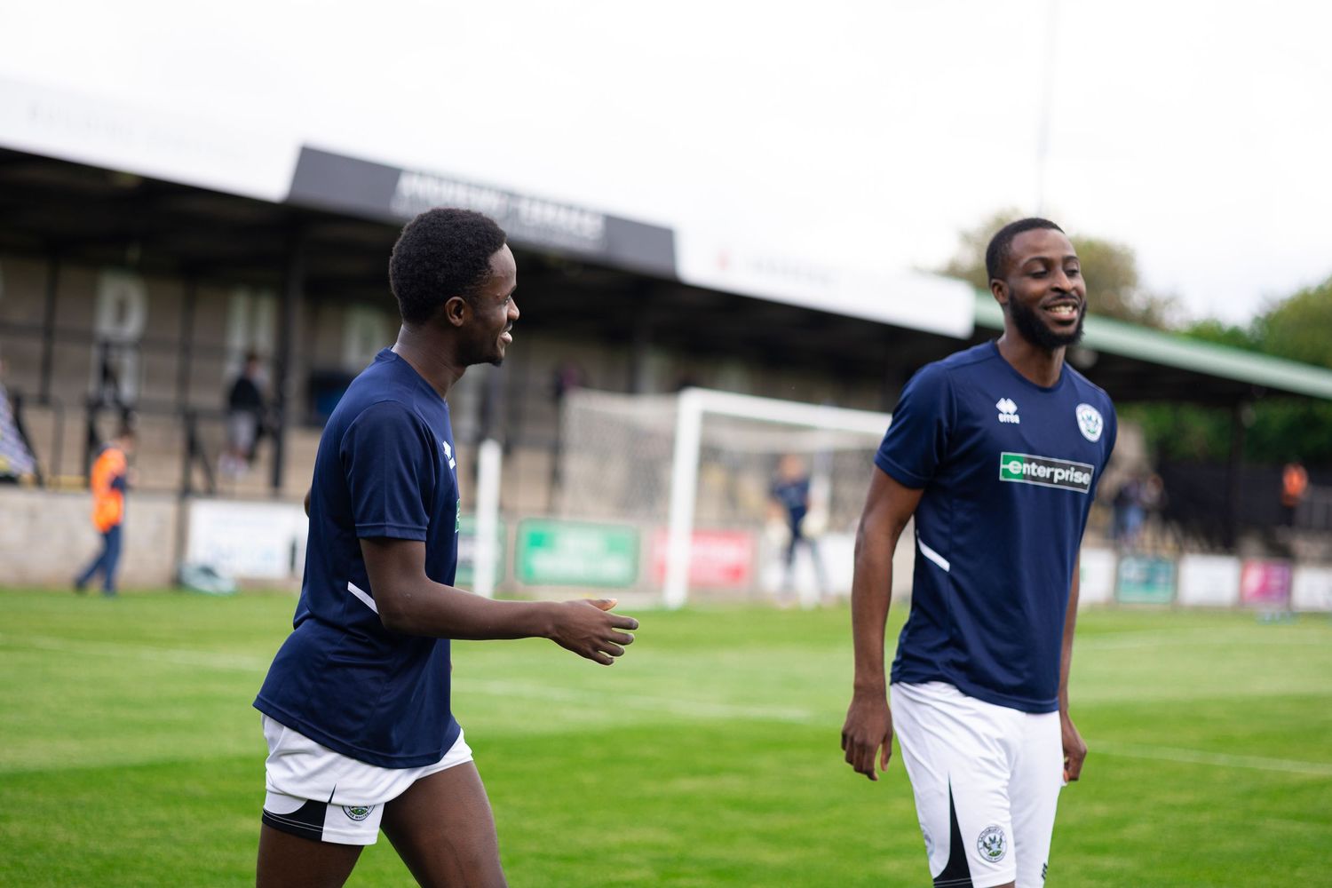 Two soccer players in navy blue uniforms laughing on a green field during a match at an outdoor stadium.