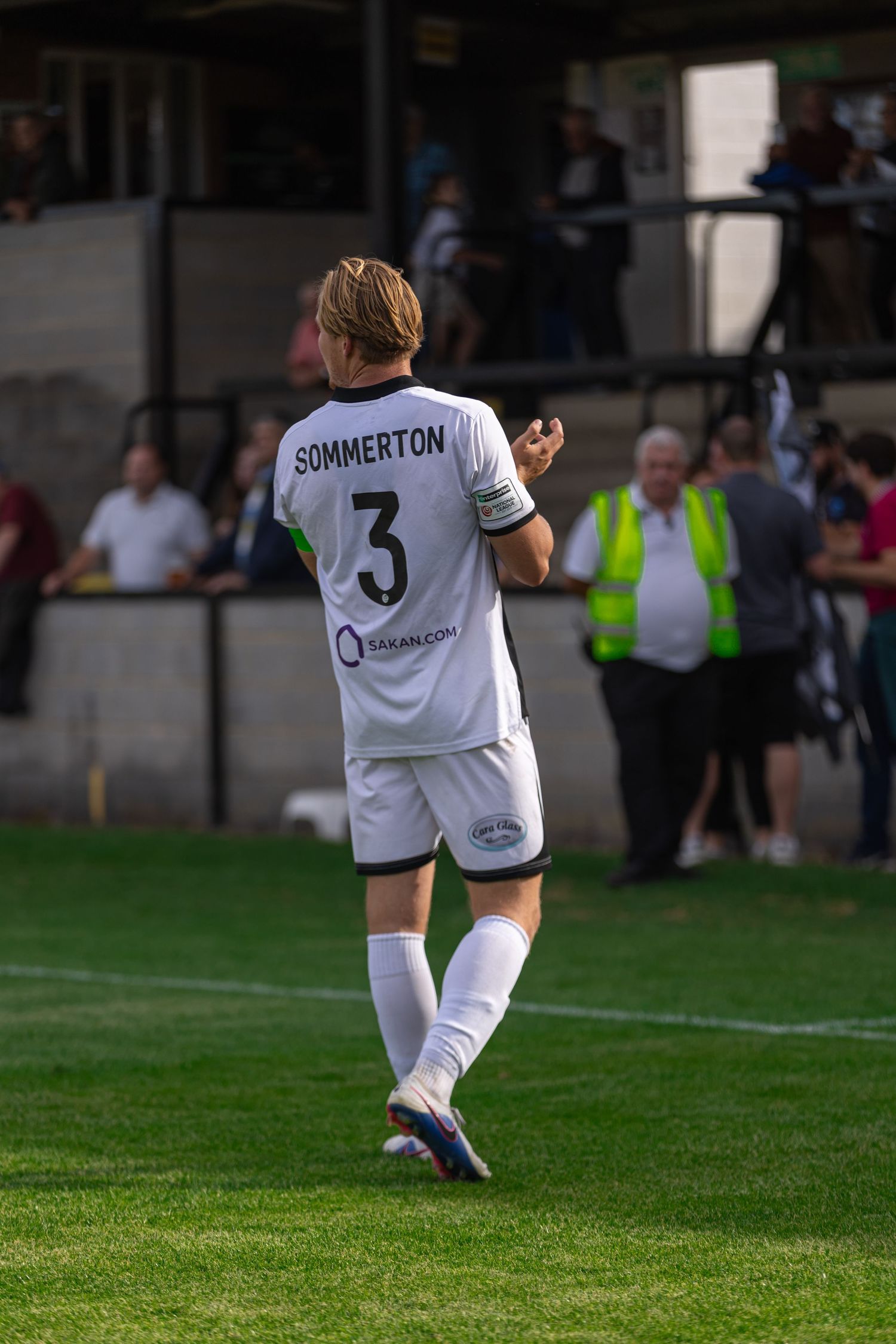 Soccer player Sommerton wearing number 3 white jersey walks on green pitch during a match with fans in background.
