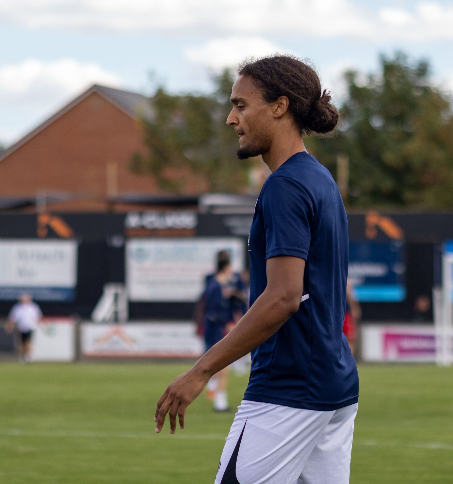 Male footballer with long hair in a navy blue kit walks across a grass pitch during pre-match warmup.
