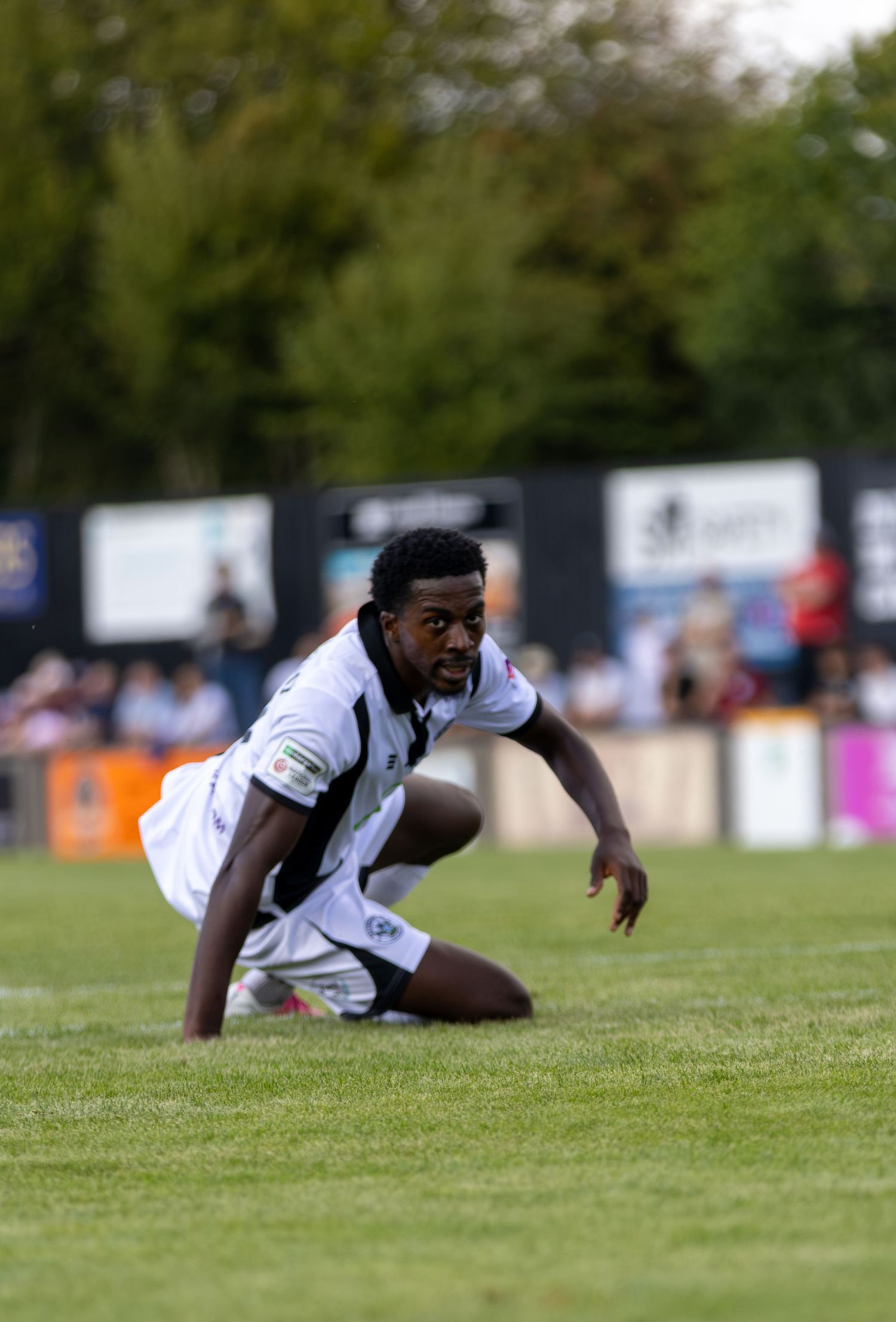 Football player in white and black kit crouches on the grass pitch during a sunny non-league match.