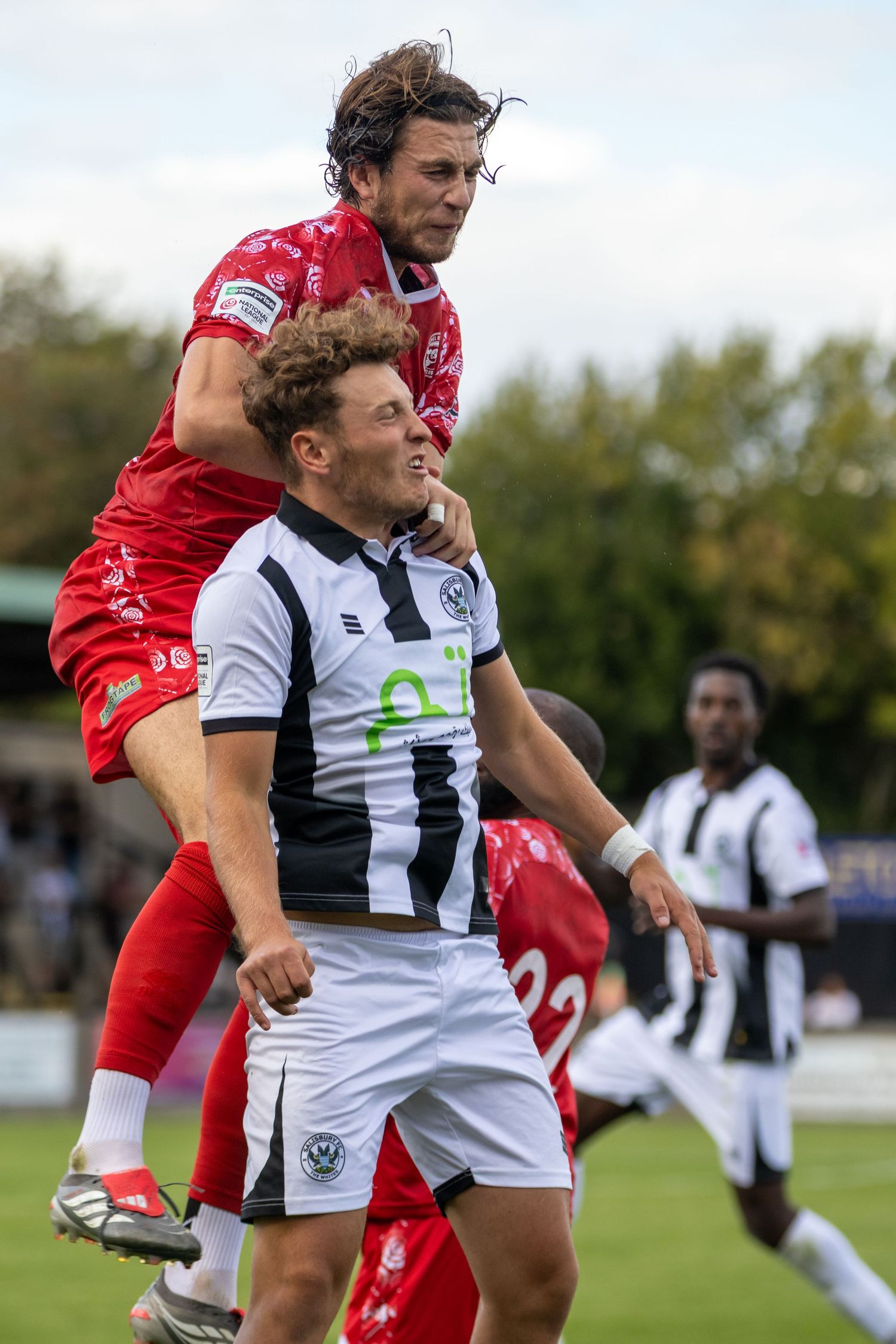 Two soccer players compete for a header during a match, one in red and one in black and white striped kit.