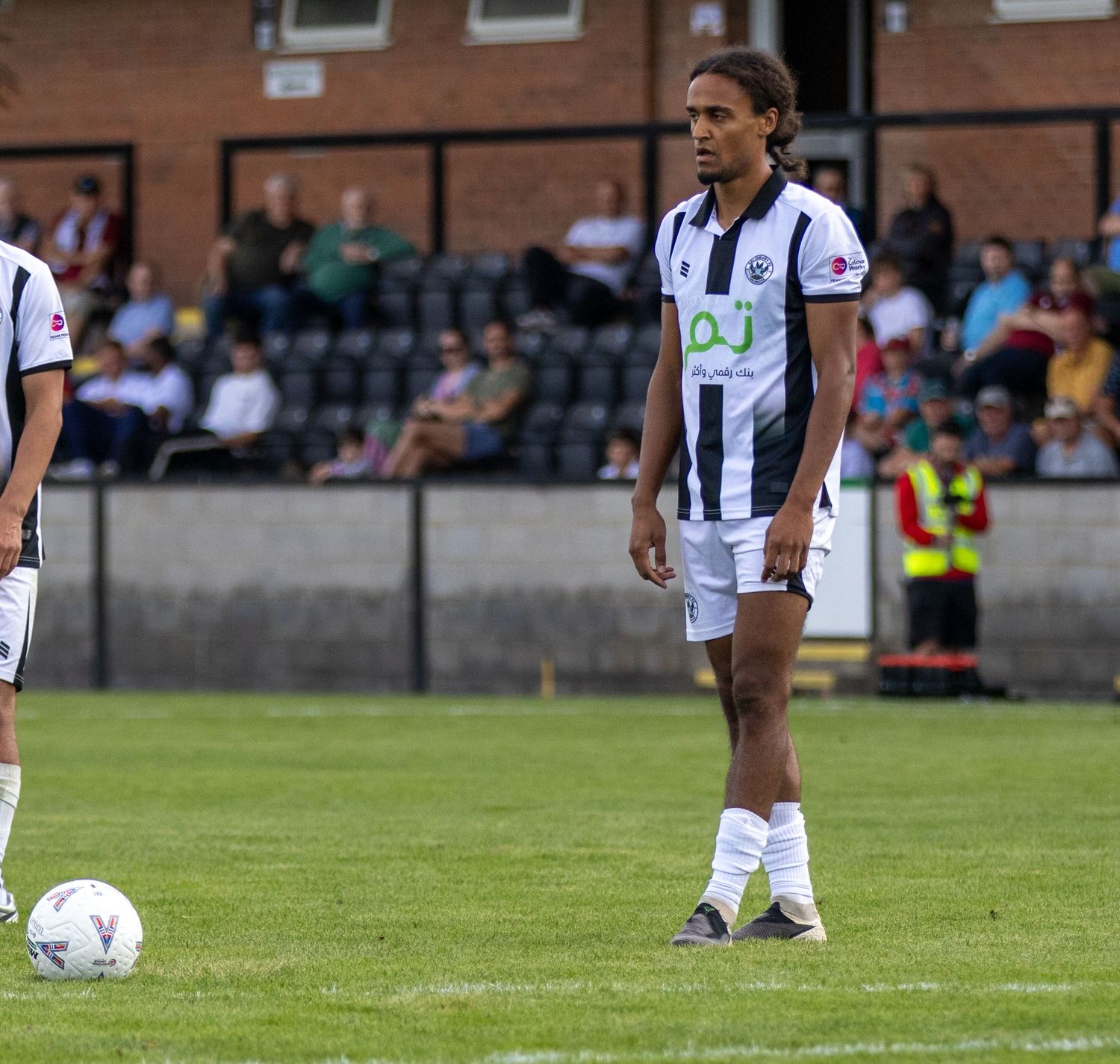 Footballer in black and white OJ-sponsored kit stands near ball on football pitch.