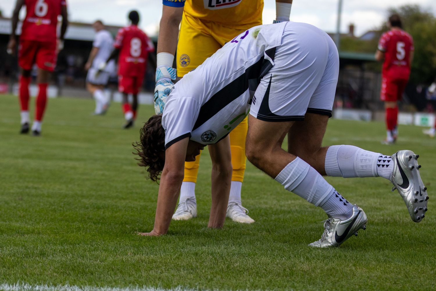 Football player in white kit bends down on the grass during a soccer match.