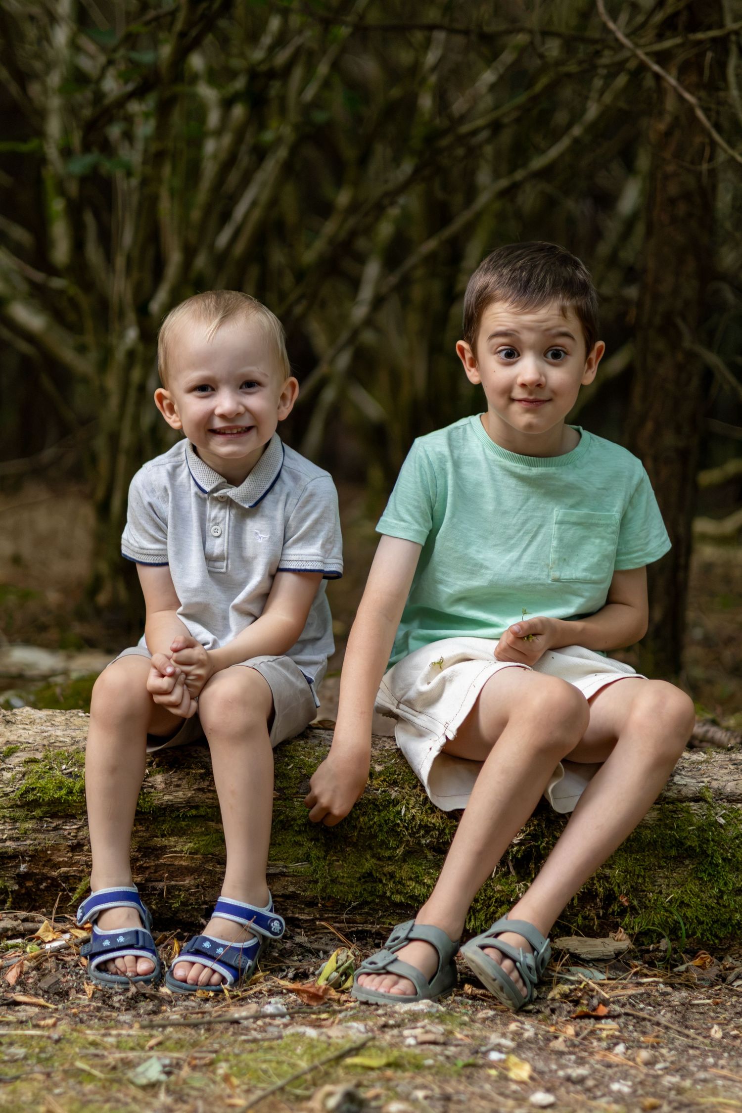 Two young brothers sit on a mossy log in a forest, smiling at the camera.