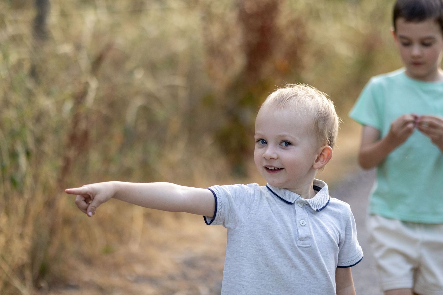 A cheerful blonde toddler in a white polo shirt pointing forward on a sunny outdoor path.