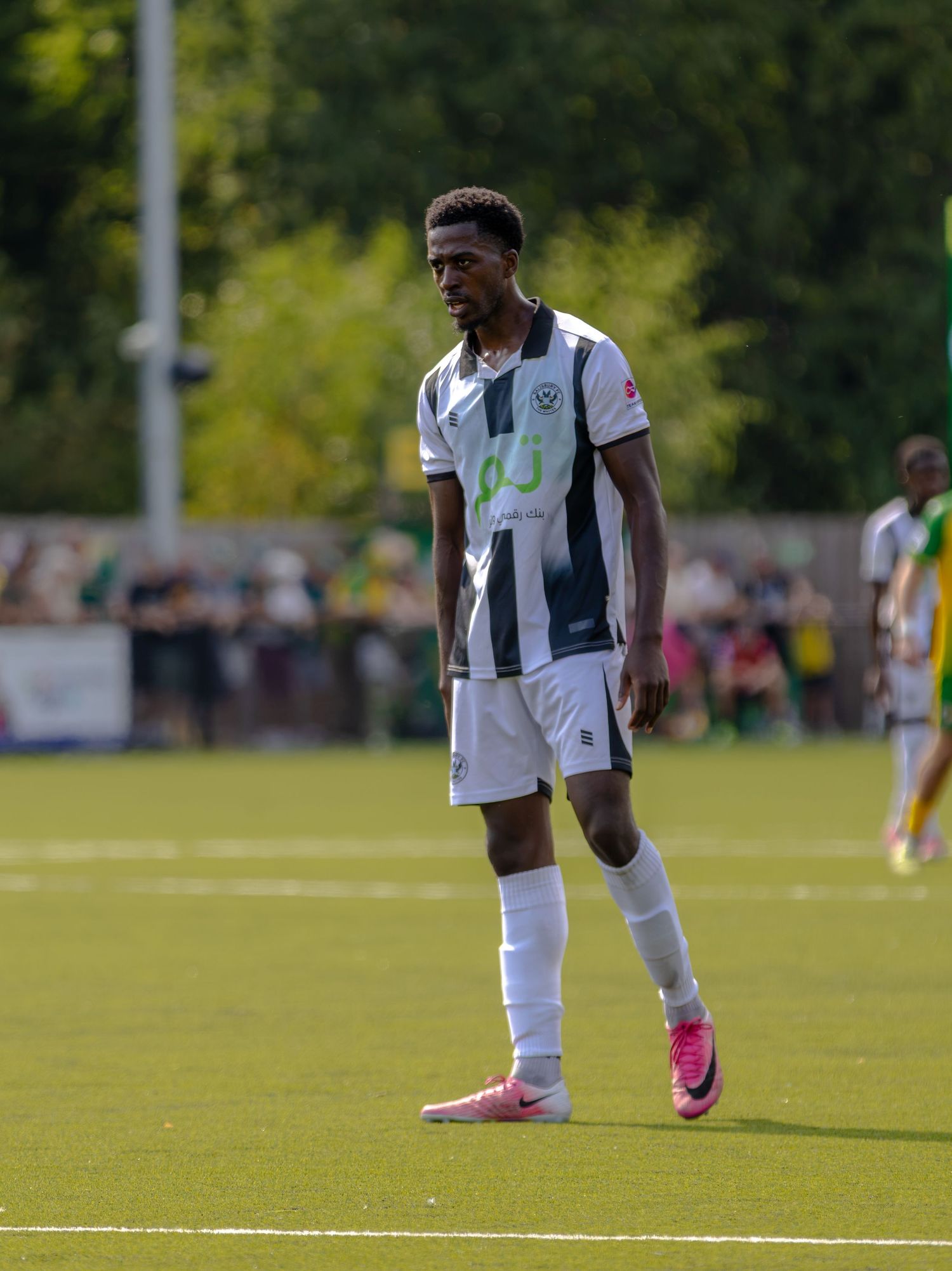 A footballer in black and white striped kit with pink boots stands on a green pitch during a sunny outdoor match.