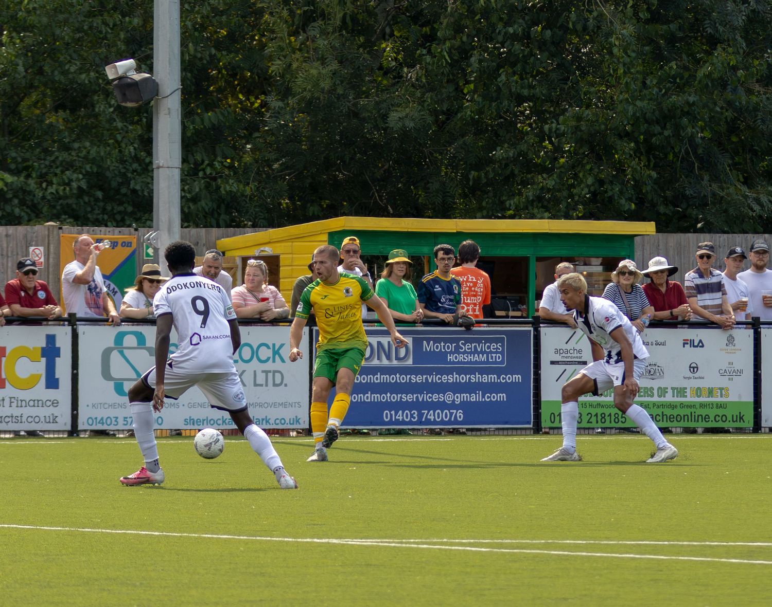 Football players in yellow-green and white kits compete for the ball on a sunny pitch.