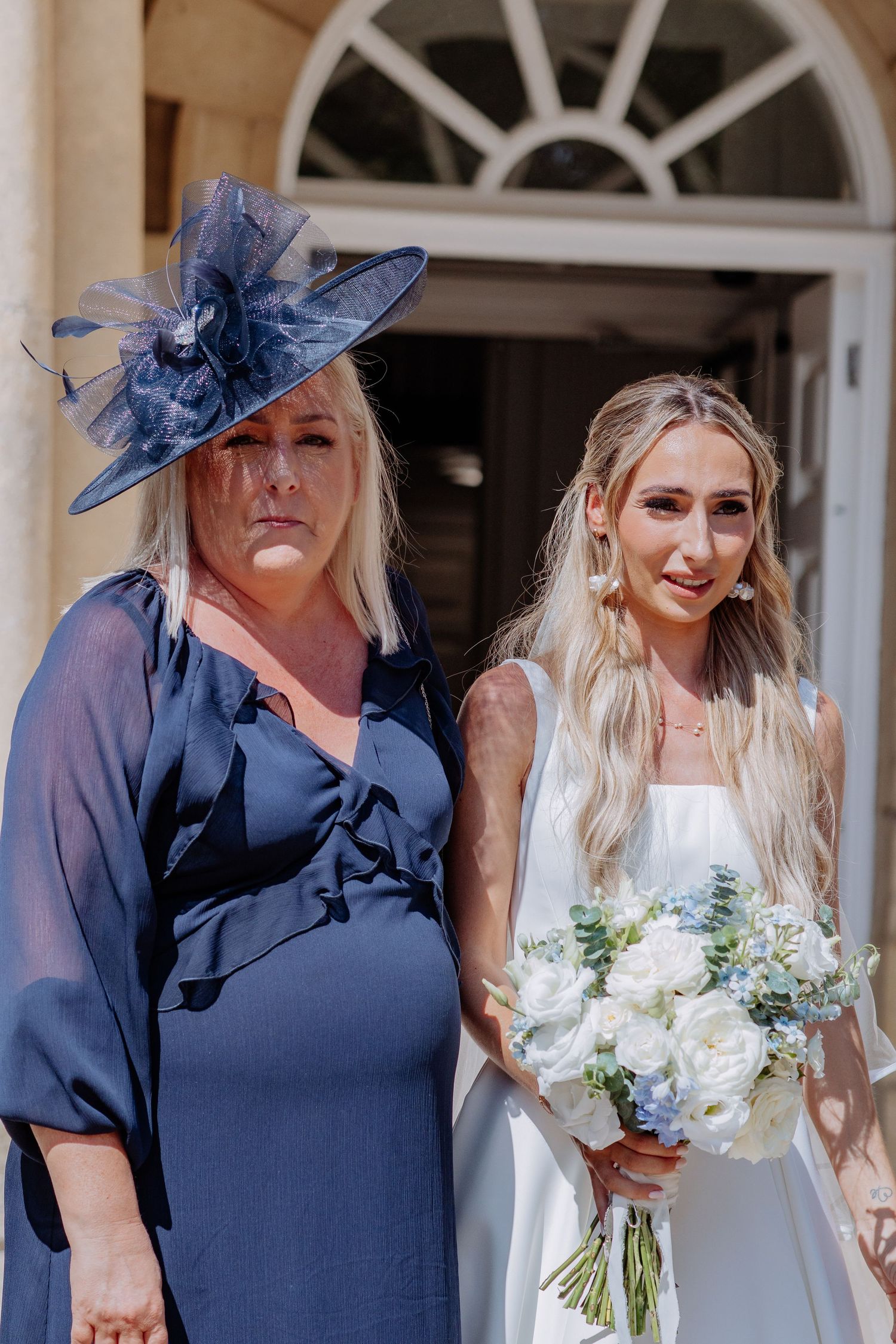 Bride holding white floral bouquet stands beside woman in navy dress and large fascinator hat at sunny wedding.