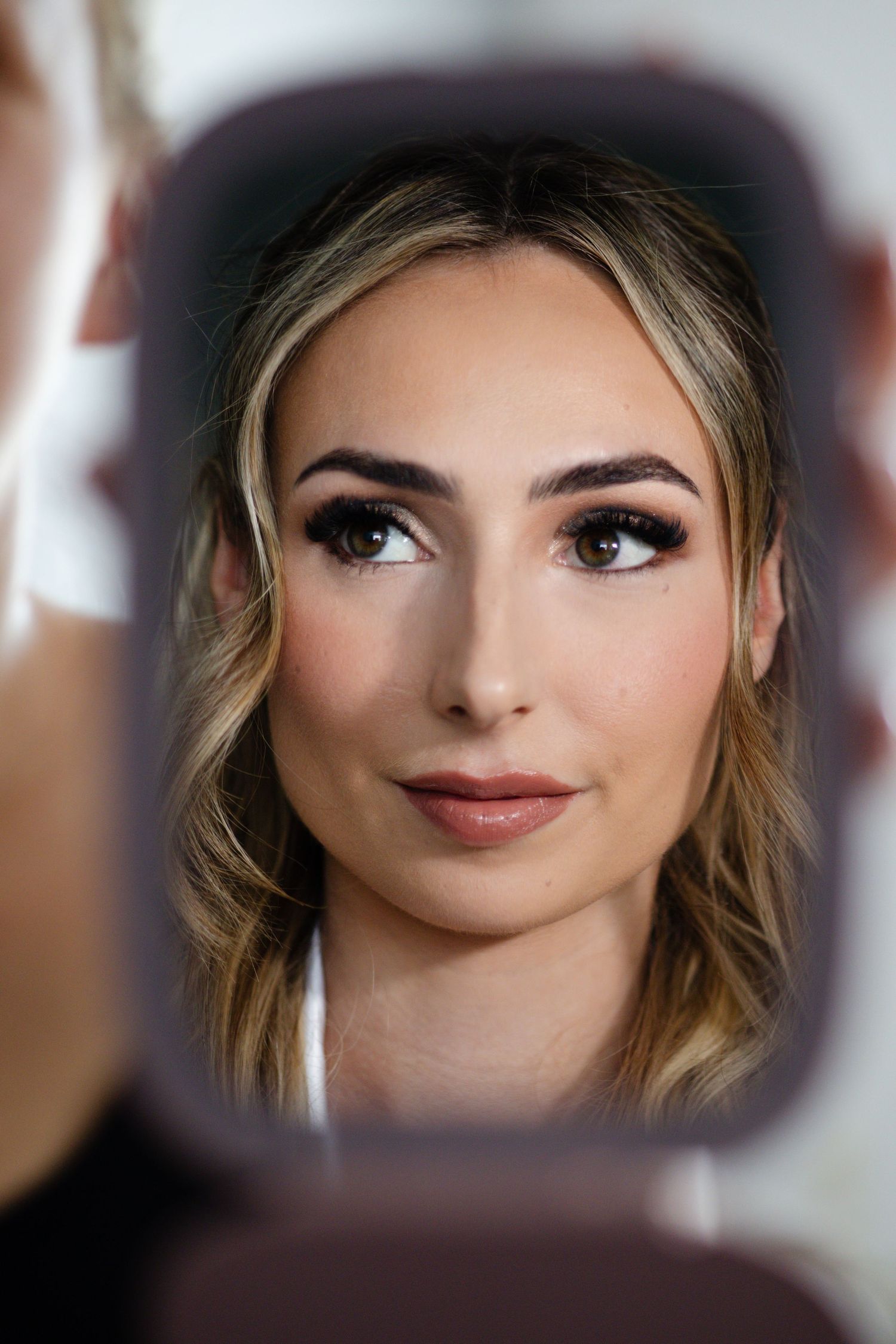 Reflection of a blonde woman with flawless bridal makeup including defined brows and nude lips in a mirror.