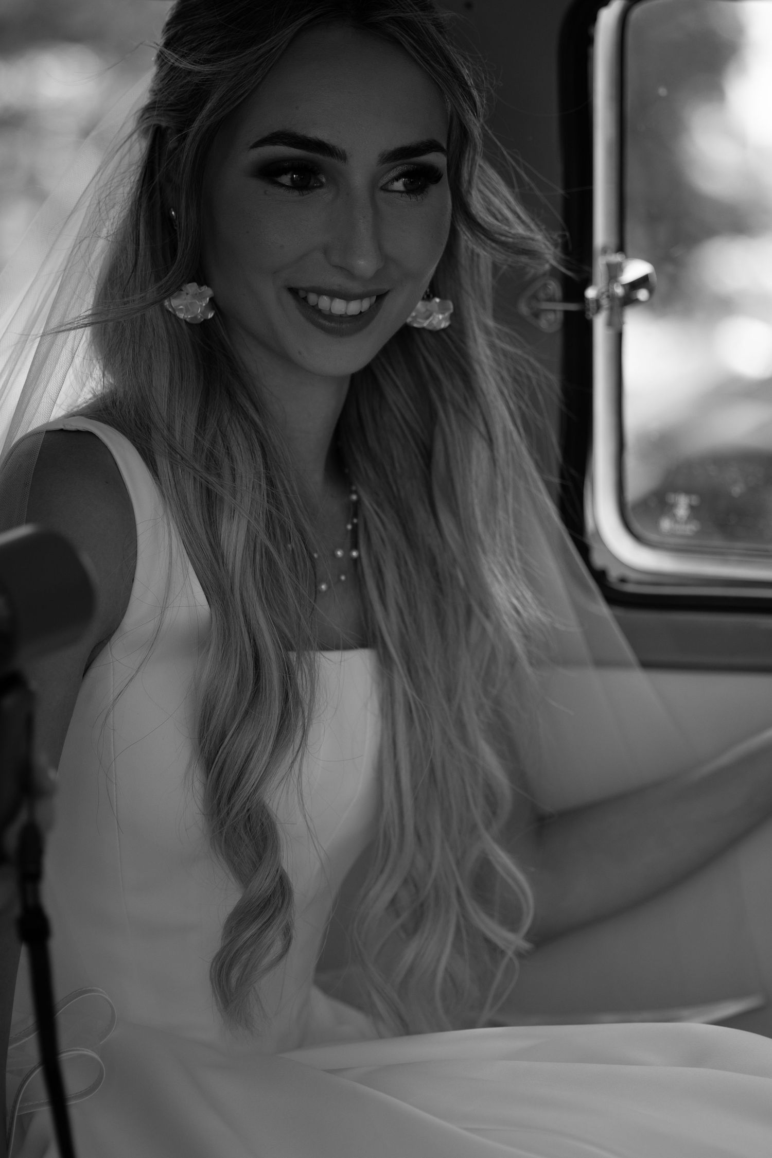Black and white portrait of a smiling blonde bride in a white gown and veil seated in a wedding car.