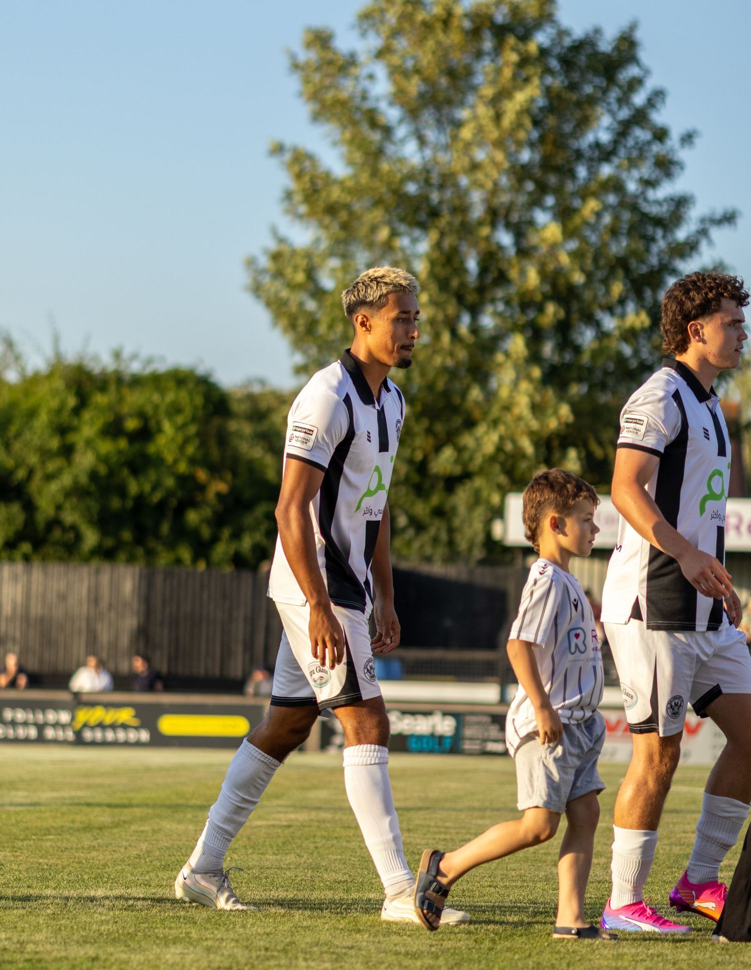 A footballer in a black and white striped kit walks onto the pitch with a young mascot on a sunny day.