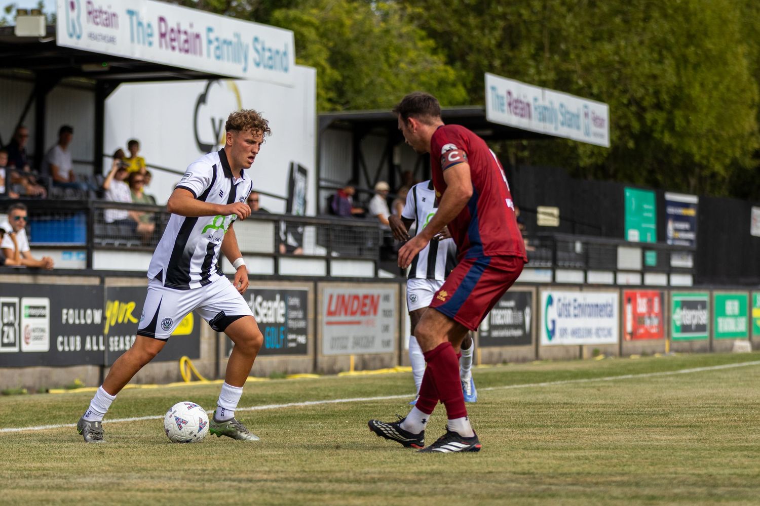 A footballer in black and white kit dribbles past a red-kitted opponent during an outdoor match on a sunny day.