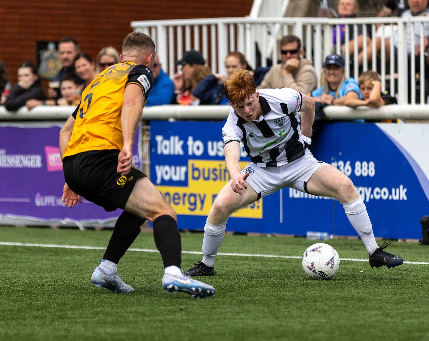 Two soccer players compete for the ball during a match, with spectators and a blue advertising board in the background.