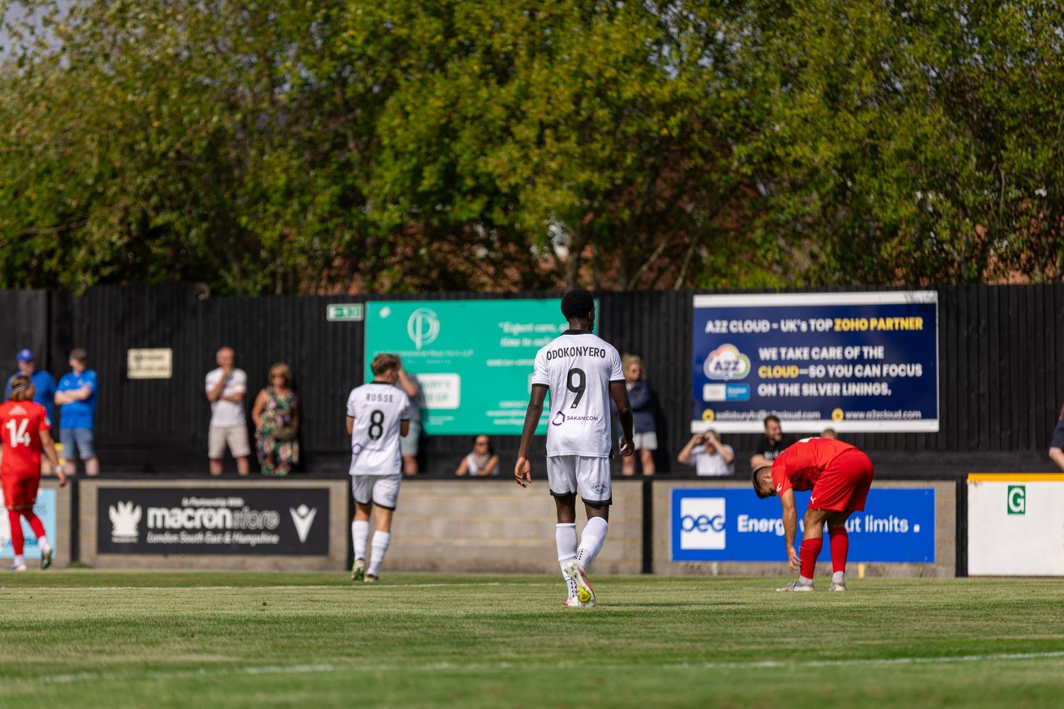 Soccer players in white jerseys numbered 8 and 9 walk on a sunny football pitch during a match.