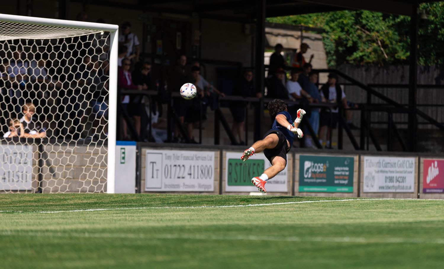 Goalkeeper diving to make a save during a sunny outdoor soccer match near the goalpost.