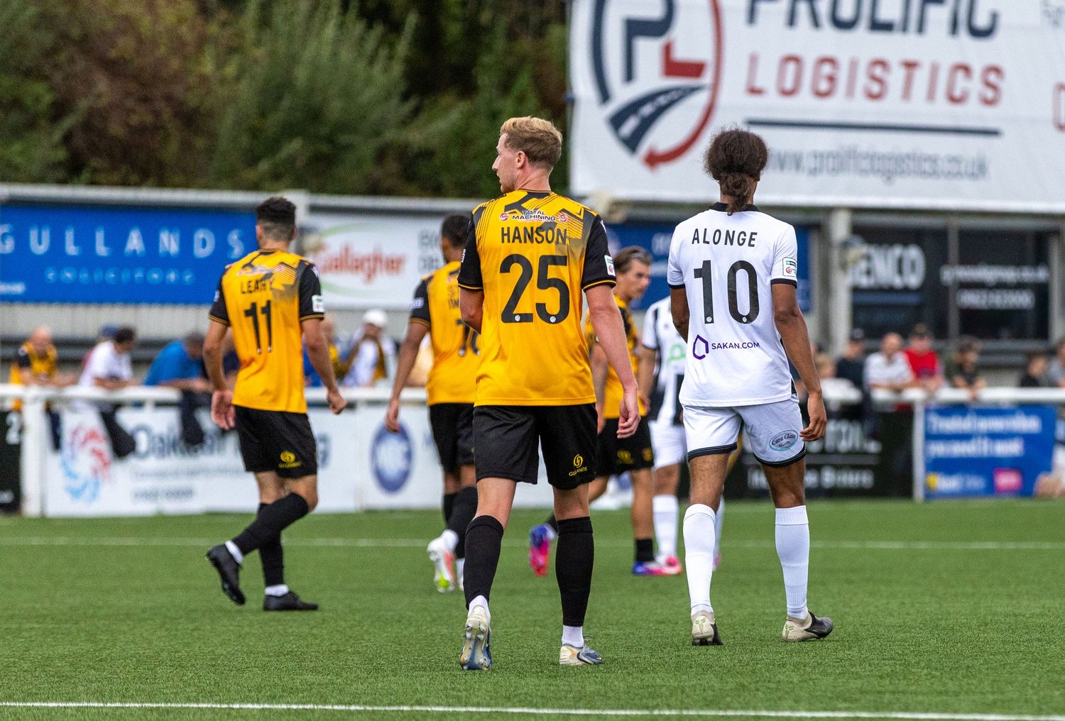 Football players in yellow and black jerseys, including number 25 Hanson, compete against white-kitted opponents on a green pitch.
